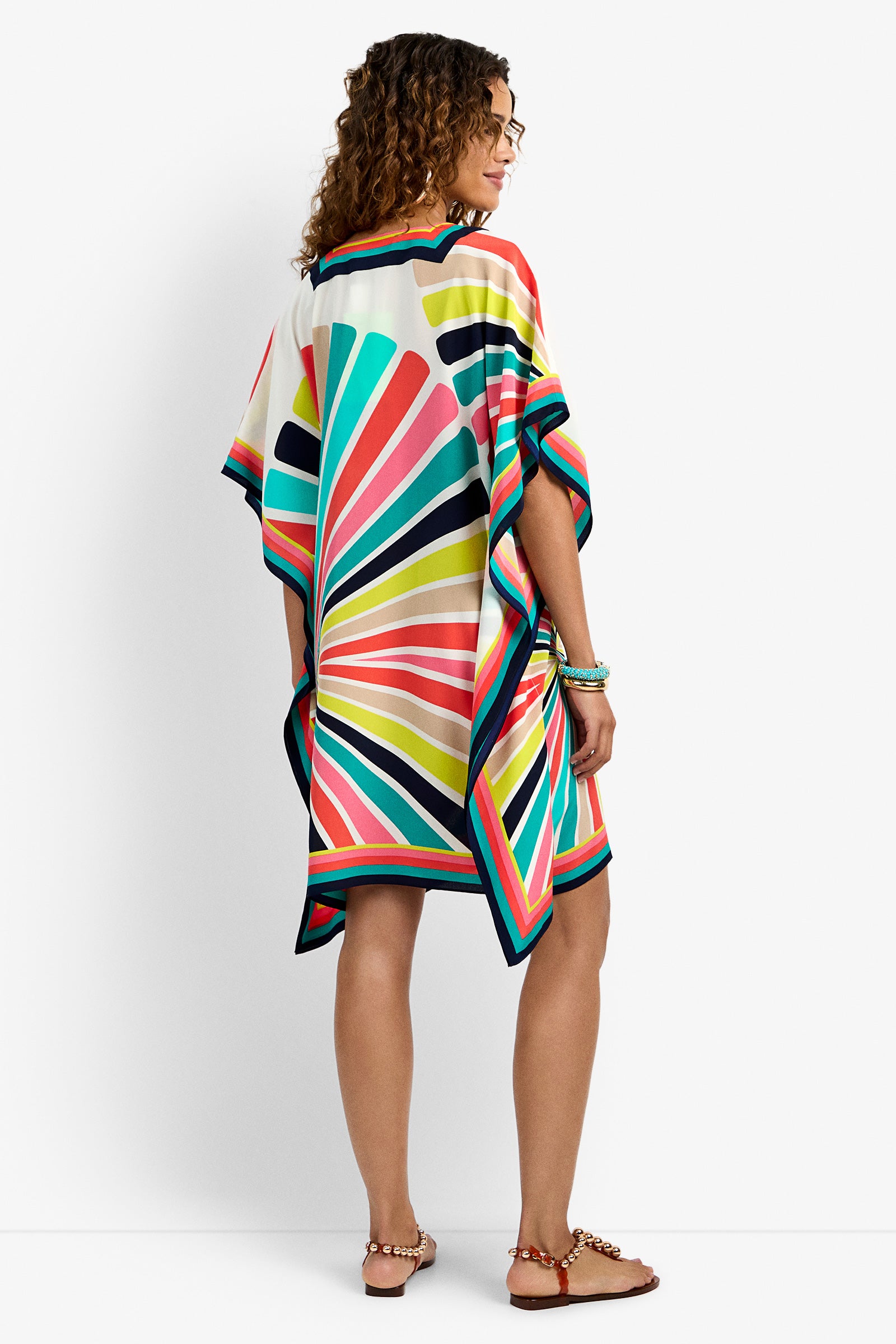 A woman with curly hair faces away, wearing the Theodora Dress—a colorful, geometric-patterned kimono-style dress—paired with sandals and bracelets, set against a plain white background.