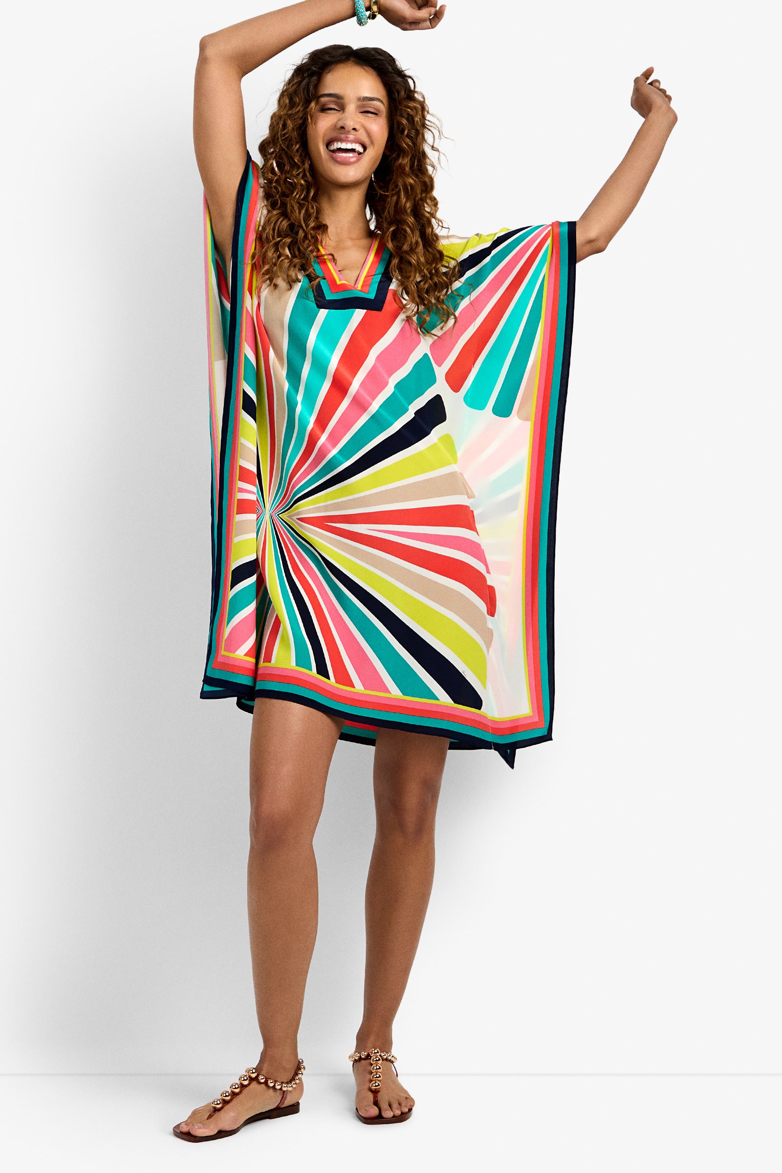A woman with curly hair smiles with arms raised, barefoot against a white background, wearing the Theodora Dress—a loose, knee-length style featuring a colorful geometric sunburst pattern.