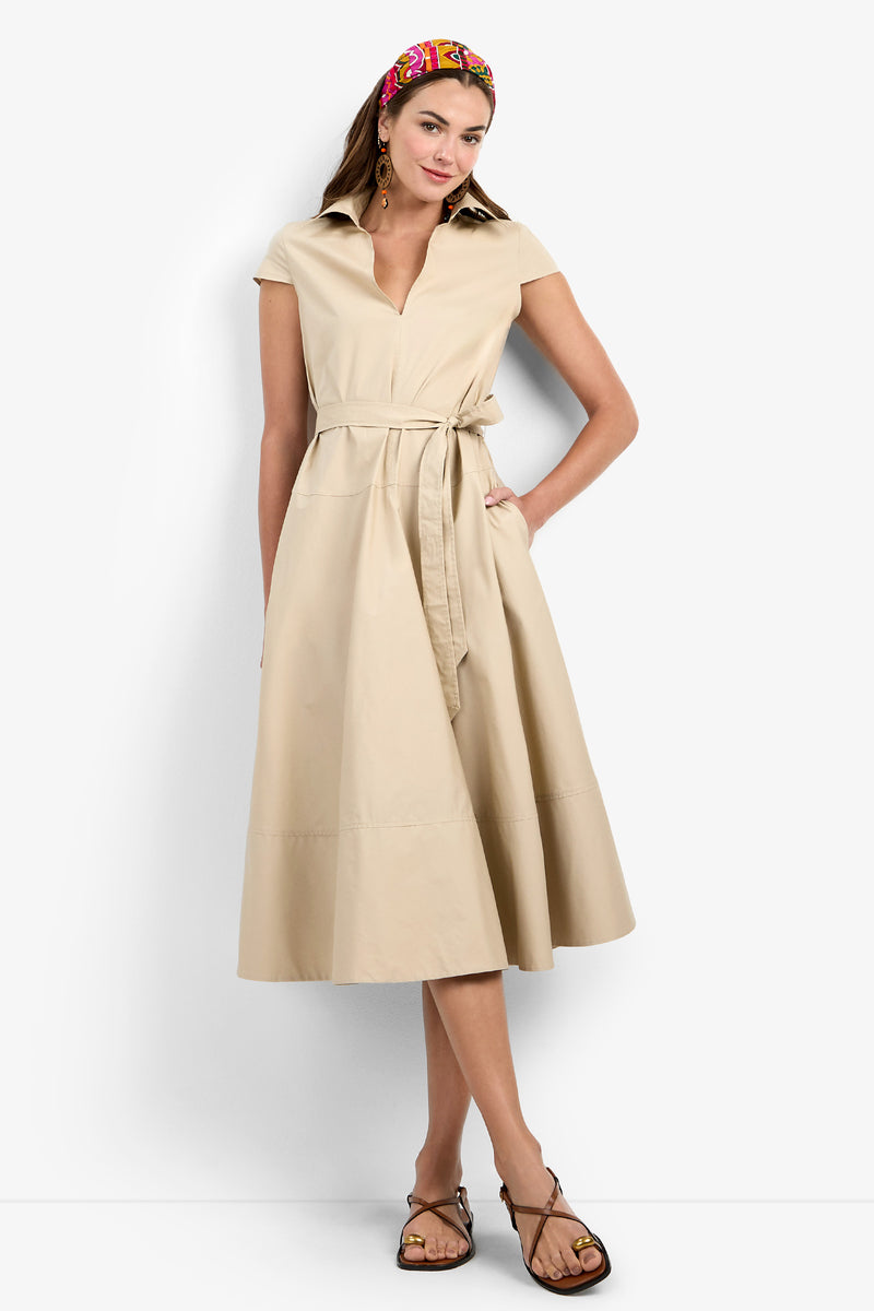 A woman wears the Delfina Dress—a beige, short-sleeve, collared midi dress with a tie belt and pockets—paired with a colorful headband, hoop earrings, and strappy sandals.