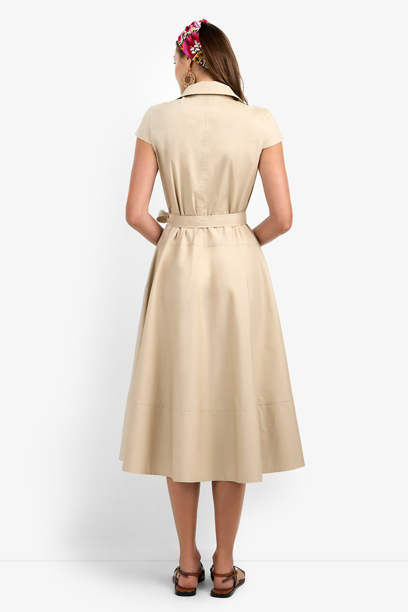 A woman faces away from the camera in a Delfina Dress—a short-sleeve beige belted midi—paired with brown sandals and a vibrant hair accessory, set against a plain white backdrop.