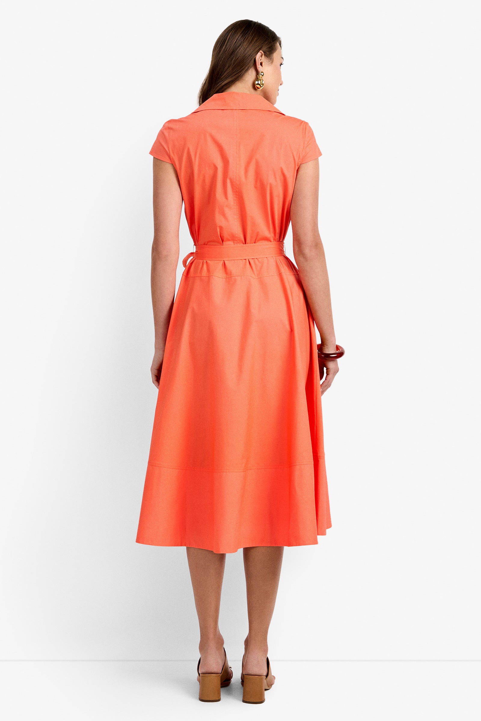 A woman with long hair faces away, wearing the DELFINA DRESS—a coral-orange, short-sleeve belted midi with a flared skirt. She pairs it with tan heeled sandals and chunky jewelry against a plain white background.