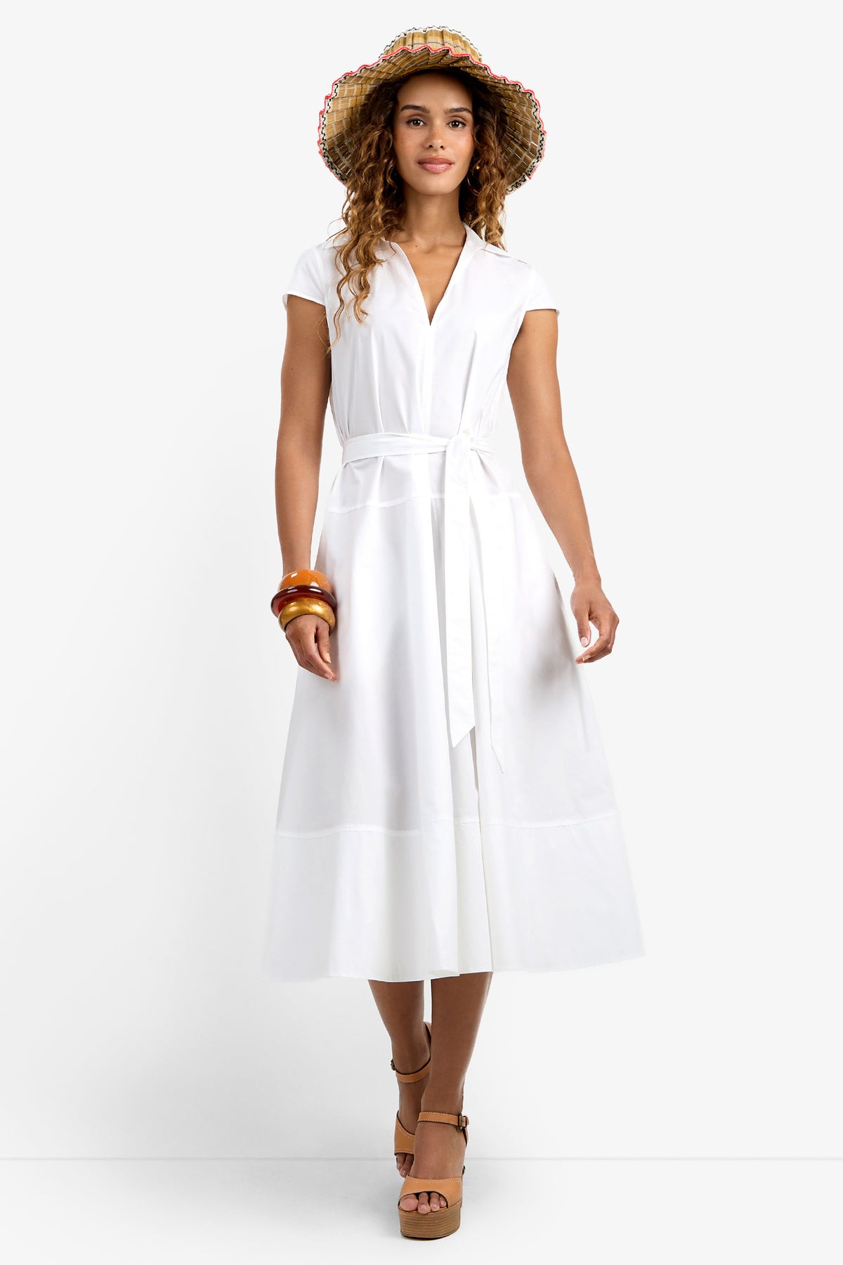 A woman in the white, short-sleeved, belted Delfina Dress, brown platform sandals, straw hat, and bracelets stands smiling against a plain white background.
