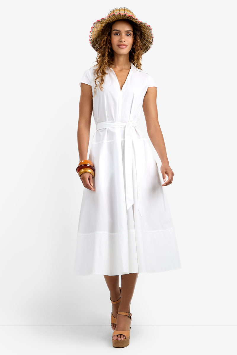 A woman in the white, short-sleeved, belted Delfina Dress, brown platform sandals, straw hat, and bracelets stands smiling against a plain white background.