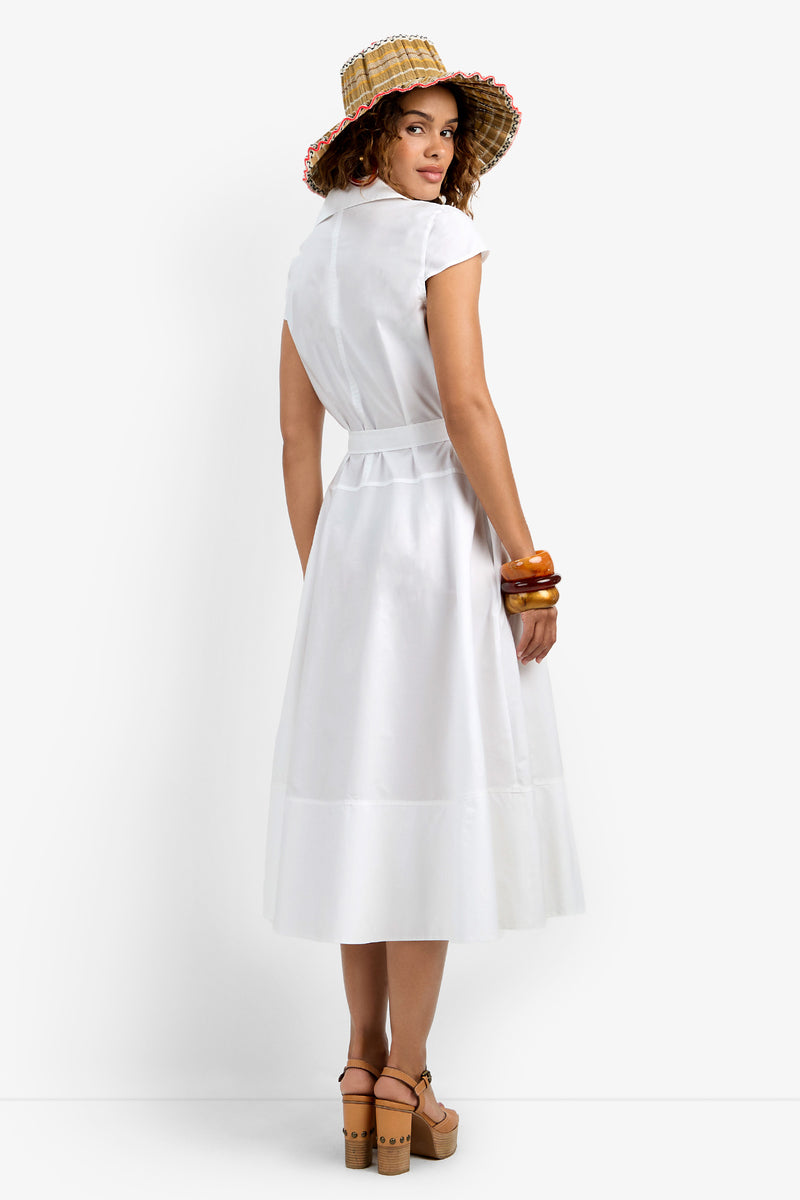 A woman in the Delfina Dress—a white, belted, short-sleeve midi—paired with a straw hat with a colorful brim, brown platform sandals, and chunky bracelets stands facing away, looking over her shoulder against a white background.