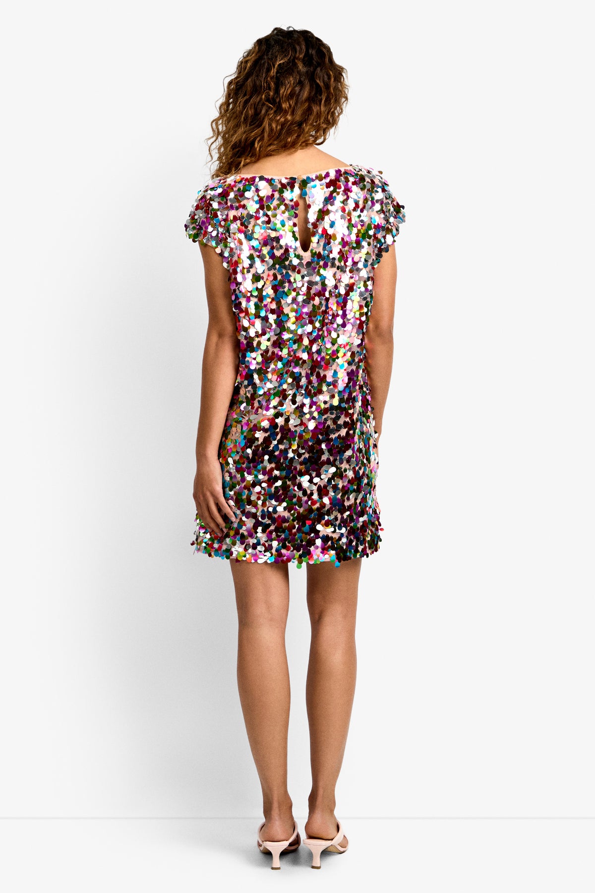 A person with curly hair is seen from the back wearing the Oceana Dress, a short multicolored sequin dress with short sleeves and a keyhole neckline, paired with light pink low-heeled shoes.