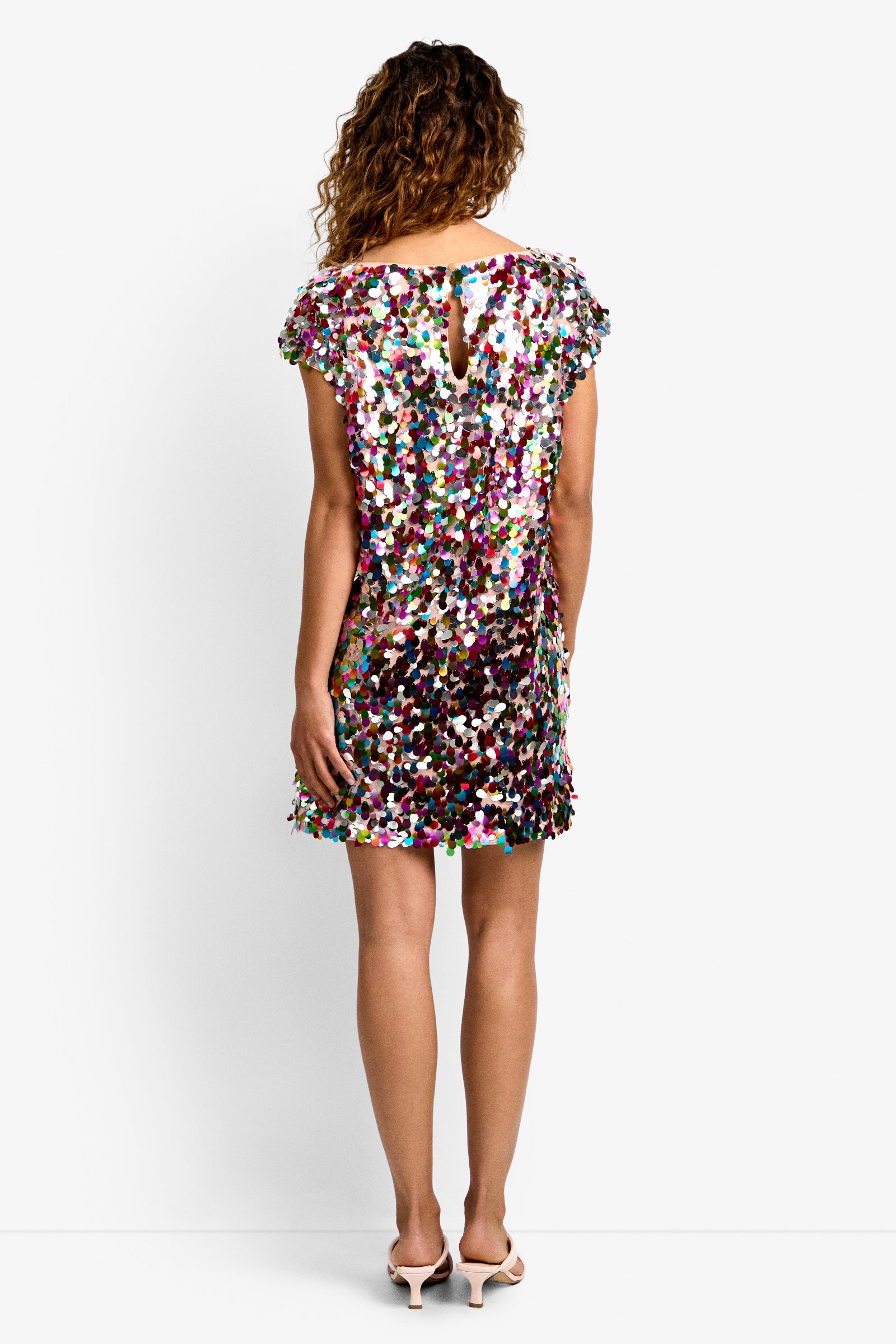 A person with curly hair is seen from the back wearing the Oceana Dress, a short multicolored sequin dress with short sleeves and a keyhole neckline, paired with light pink low-heeled shoes.