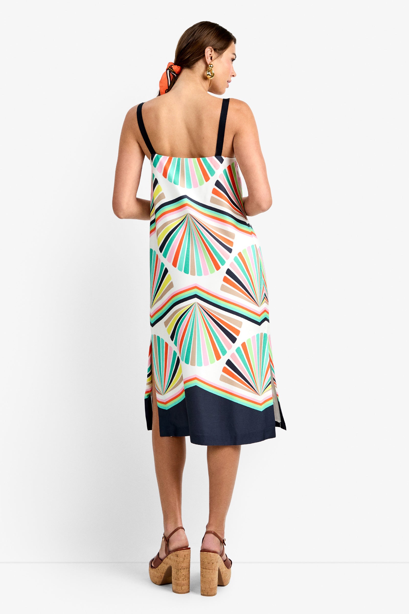 Woman with her back to the camera wears the Bayside Dress, a colorful sleeveless geometric-patterned piece with navy straps and hem, paired with wooden platform sandals, gold earrings, and an orange hair scarf.
