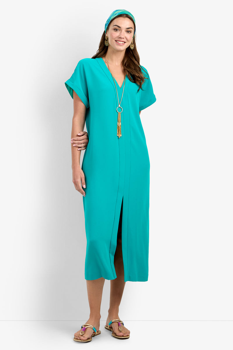 A smiling woman wears the OASIS DRESS, a teal midi dress with short sleeves and a front slit, paired with a matching headband, long necklace, hoop earrings, and sandals against a white background.