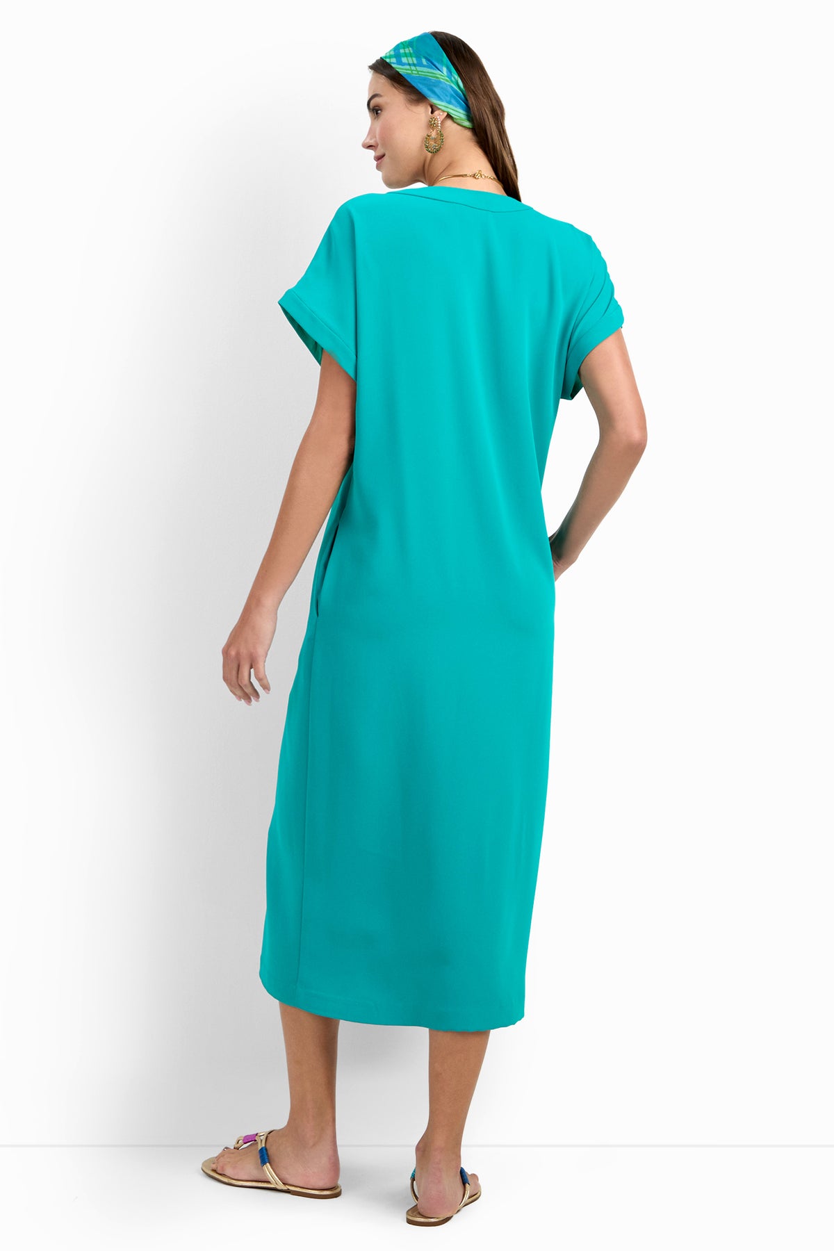 A woman stands with her back to the camera, wearing the OASIS DRESS—a loose-fitting, knee-length turquoise dress—with flat sandals and a matching headscarf, set against a plain white background.