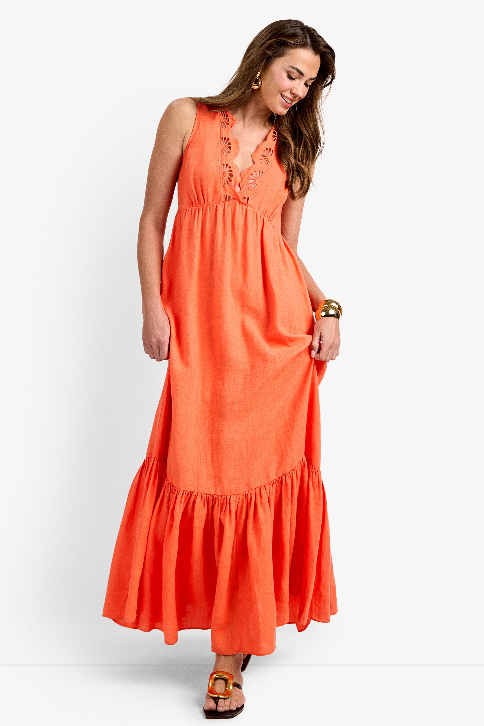 A woman smiles as she looks down, wearing the Lobster Dress in CALYPSO CORAL—a sleeveless maxi with a ruffled hem and embroidered V-neckline. She completes her look with gold earrings, bangles, and brown sandals.