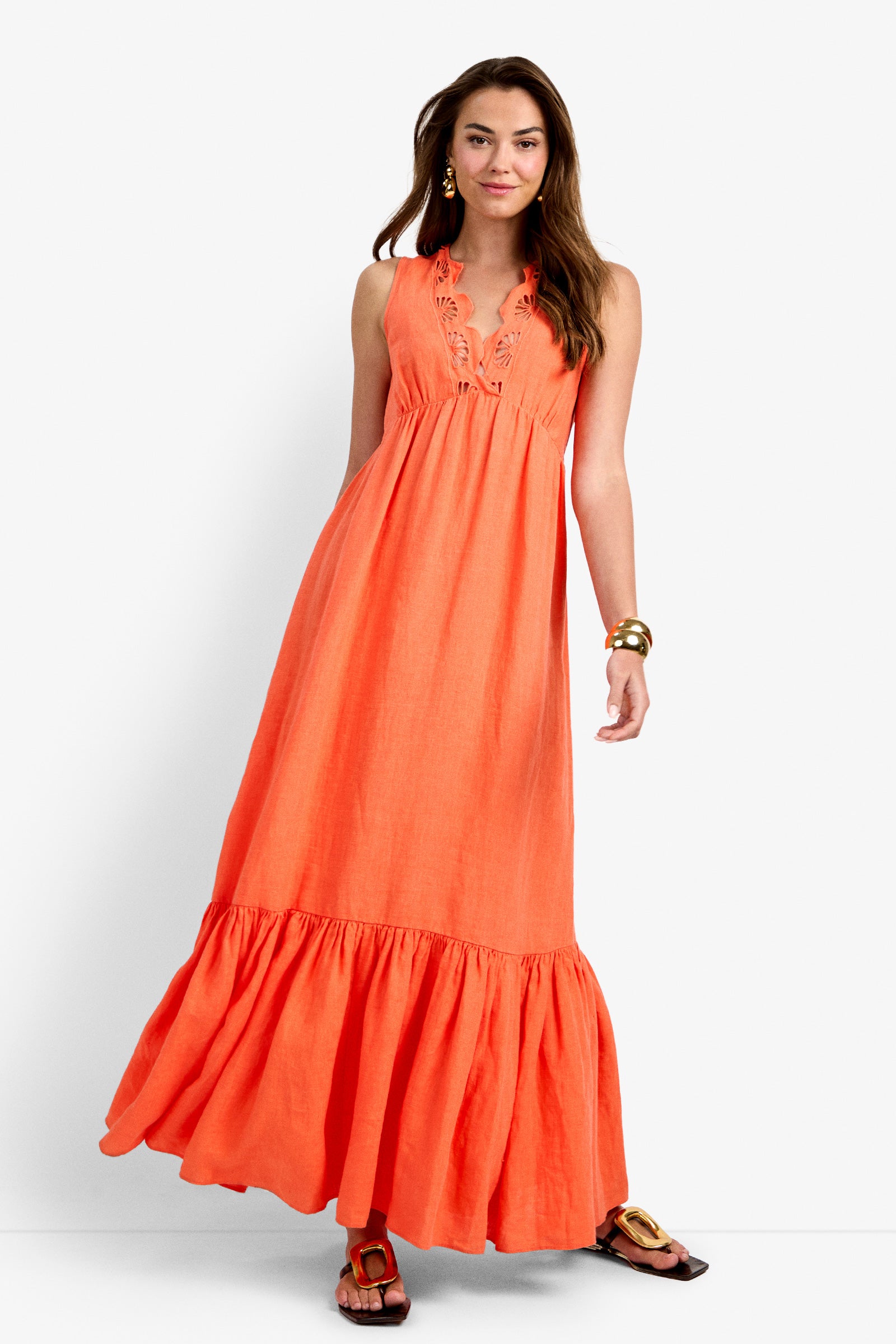 A woman wearing the Lobster Dress—a sleeveless, long orange sundress with a ruffled hem and detailed neckline—paired with sandals, stands against a plain white background.