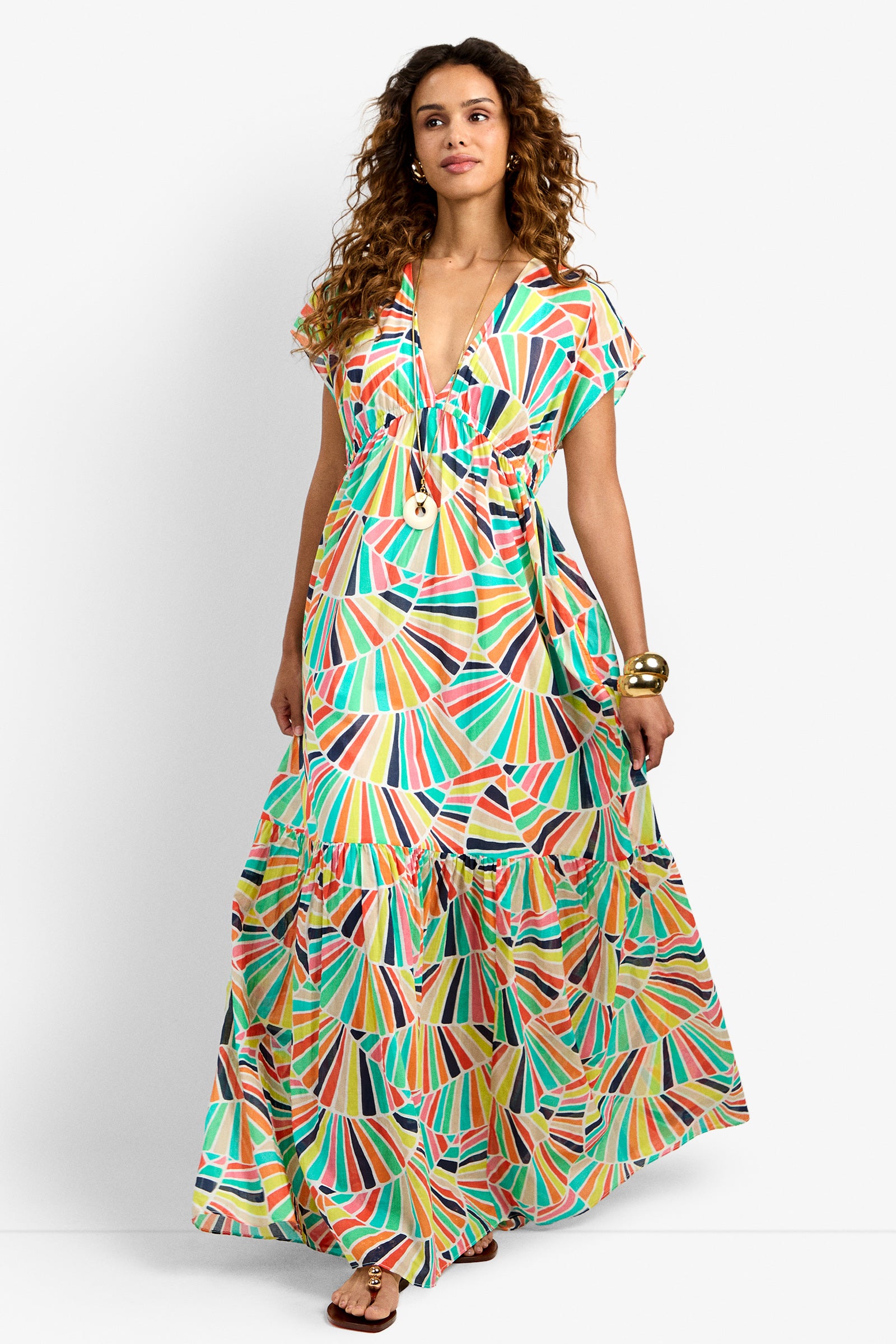Woman wearing the CAPECOD DRESS, a colorful patterned maxi with a V-neck and short sleeves, styled with gold bangles and sandals, stands against a plain white background.