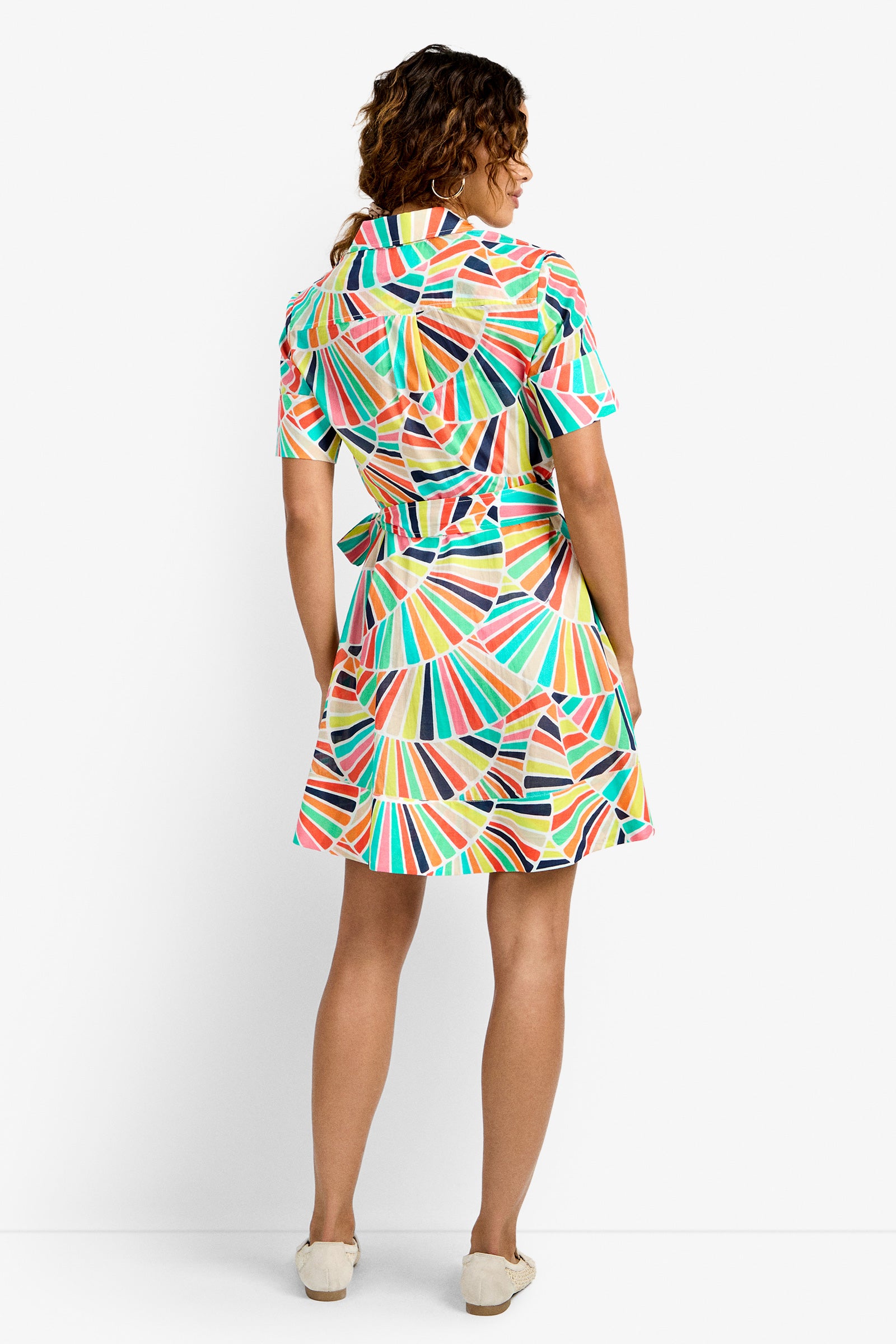 A woman faces away, wearing the Misola Dress—a colorful, short-sleeved dress with a geometric rainbow pattern and a waist belt—paired with beige slip-on shoes against a plain white background.