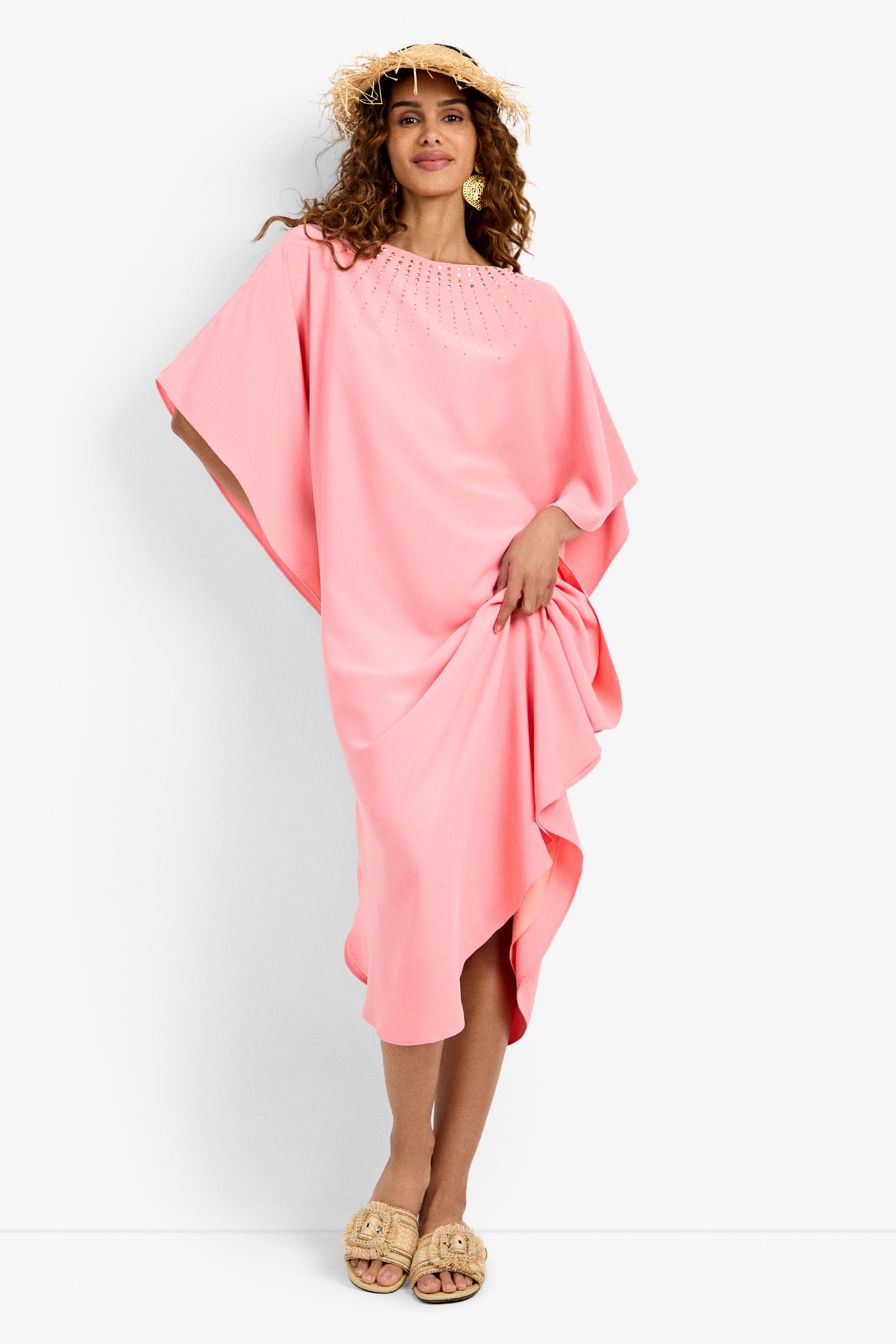 A woman in the Neena Rhinestone Caftan Dress, a loose-fitting light pink style with beaded details, stands against a white background wearing a straw hat and tan woven sandals, smiling while holding her dress to the side.