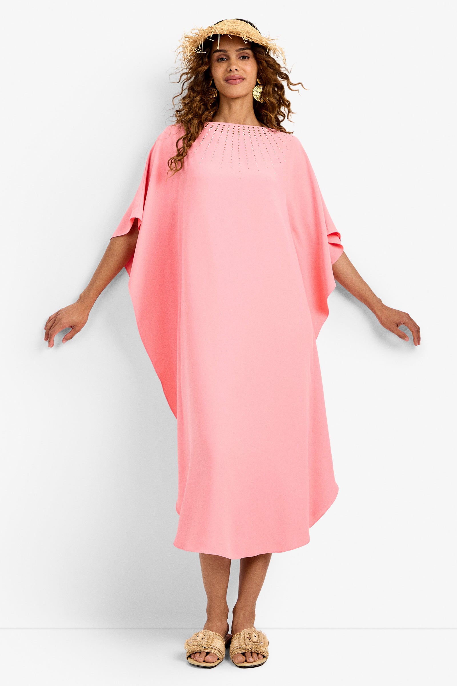 A woman stands against a white background, arms slightly extended, wearing the Neena Rhinestone Caftan Dress—a loose light pink dress with short wide sleeves—paired with a straw hat and beige sandals.