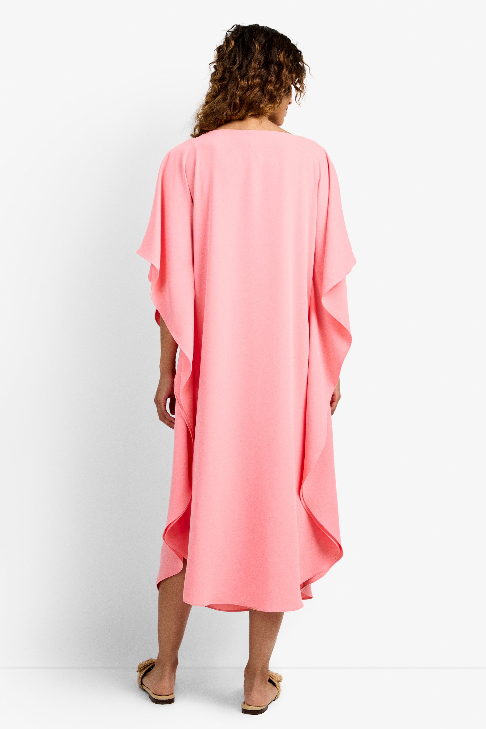 A woman with curly hair, seen from behind, wears the Neena Rhinestone Caftan Dress—a loose, flowing bright pink knee-length dress with draped sleeves—paired with tan flat sandals against a plain white background.
