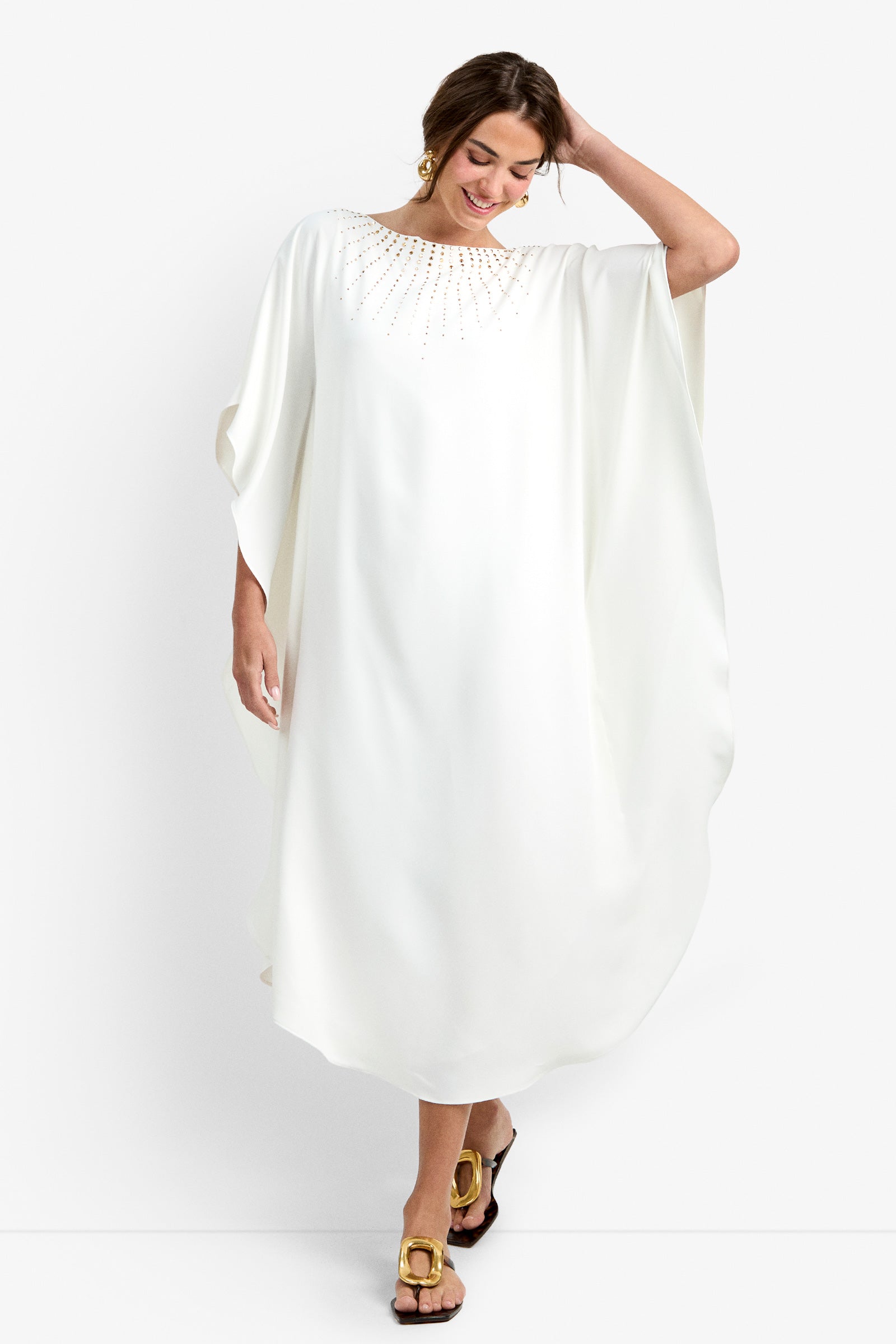A woman in the Neena Rhinestone Caftan Dress, featuring a loose white silhouette with gold embellishments at the neckline, accessorized with gold earrings and sandals, stands smiling against a white background, one hand touching her hair.