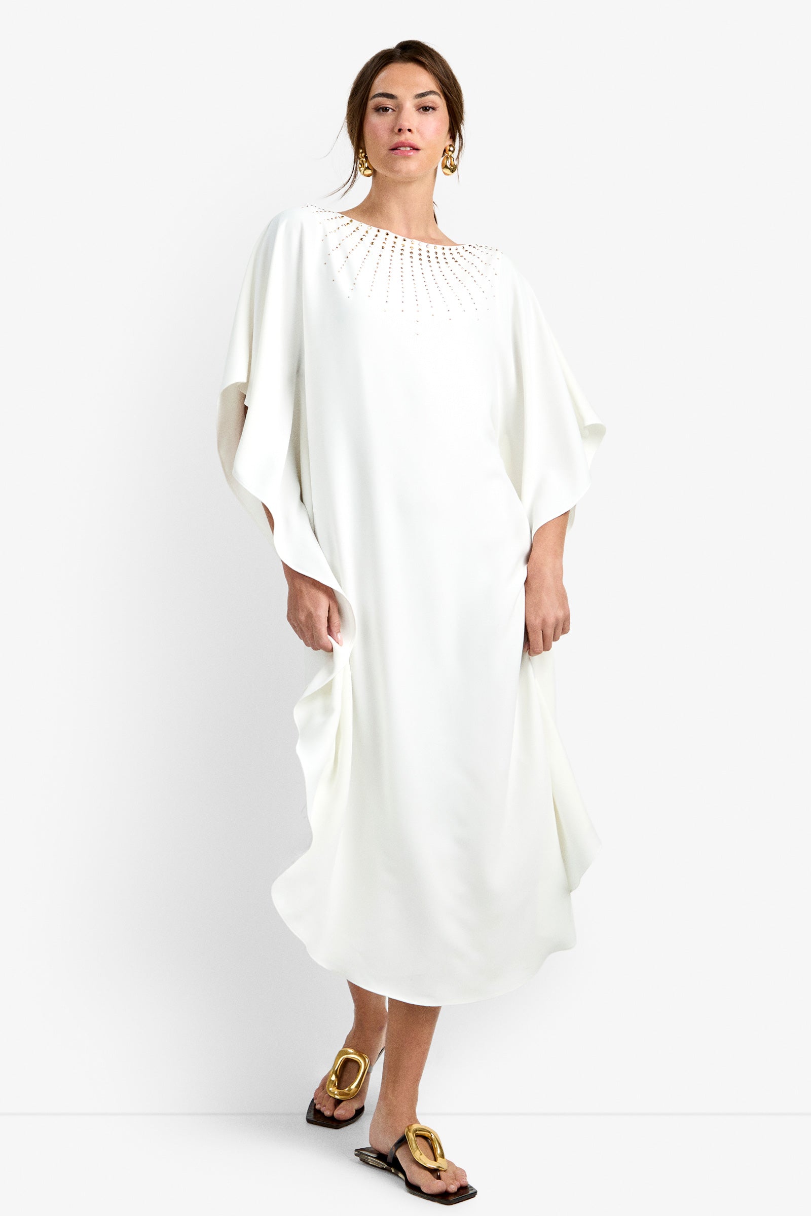 A person wearing the Neena Rhinestone Caftan Dress, gold drop earrings, and gold flat sandals stands against a plain white background.