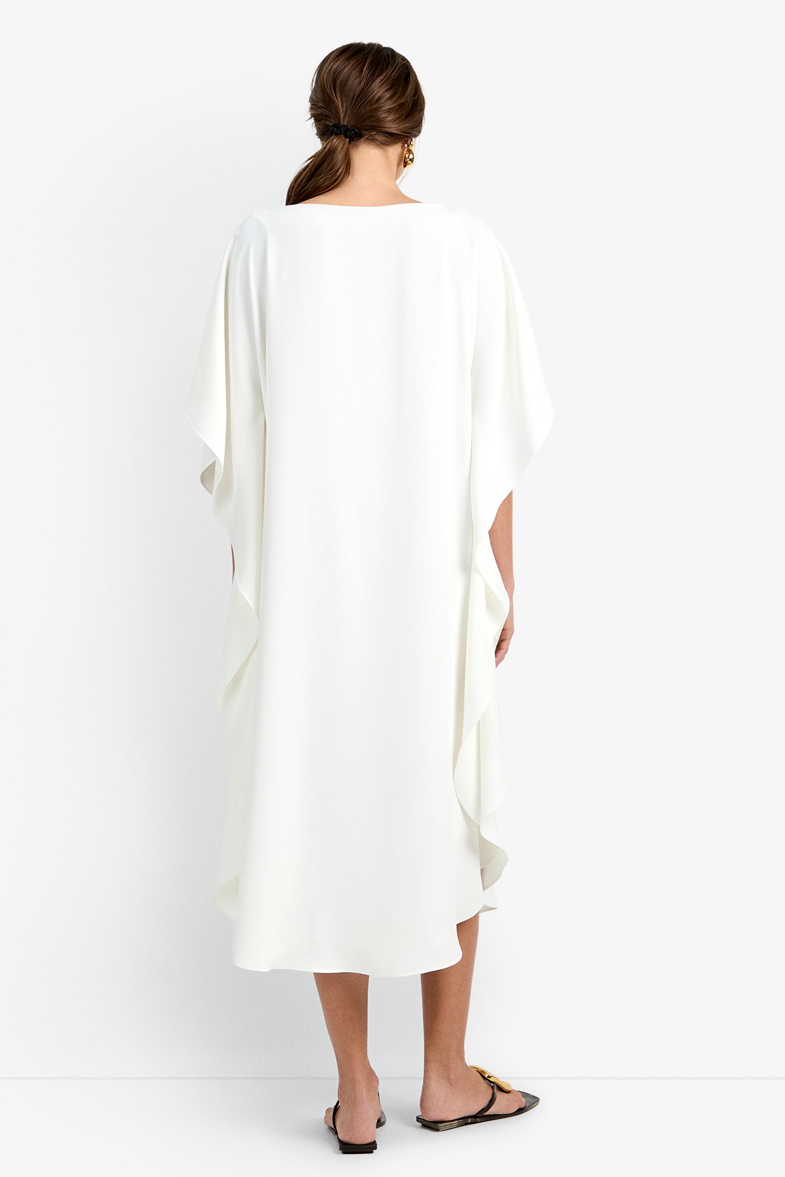 A woman faces away, wearing the Neena Rhinestone Caftan Dress—loose, white, knee-length with draped sleeves—and black flat sandals. Her brown hair is in a low side braid against a plain white background.