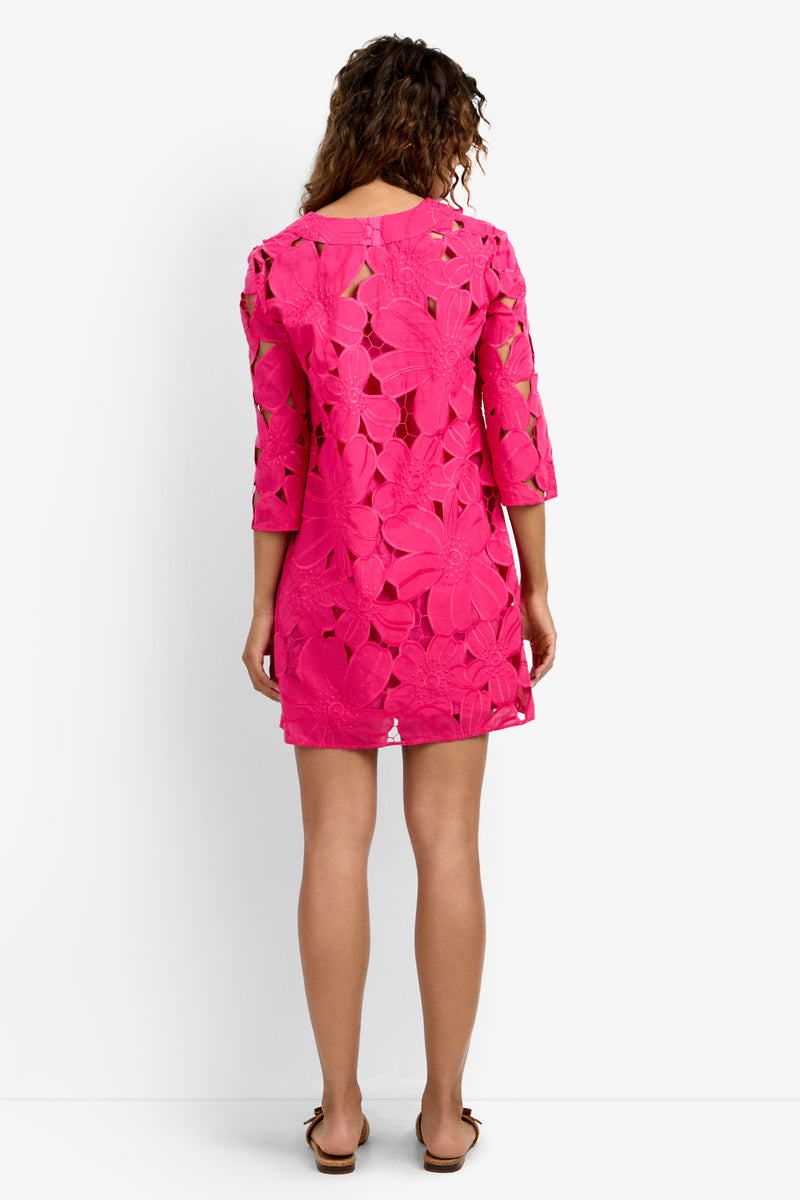 A woman with curly hair faces away, wearing the Mangrove Dress—a bright pink, knee-length dress with a floral cut-out pattern and three-quarter sleeves—paired with brown sandals against a plain white background.