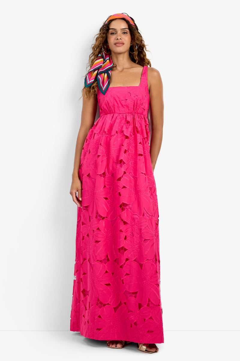 A woman in the Ariel Dress—a bright pink, sleeveless, floor-length dress with large floral cutouts—paired with a colorful headscarf and sandals, standing against a plain white background.