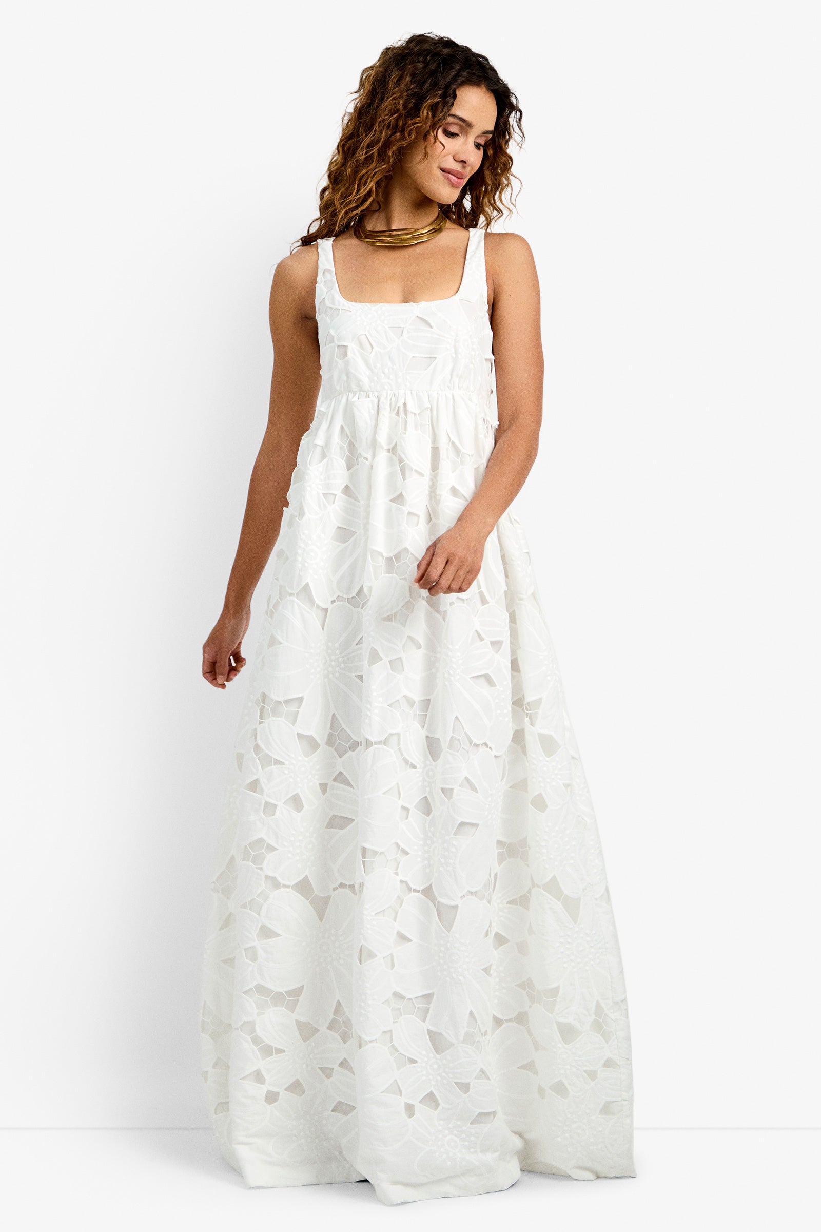 A woman stands against a white background, gently smiling to the side as she wears the Ariel Dress in WHITEWASH—a sleeveless, floor-length style featuring bold floral cut-out patterns.
