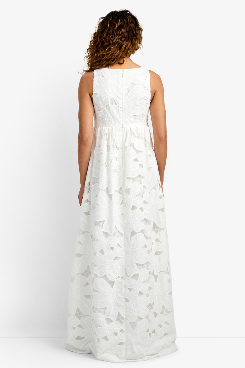 A woman with curly hair faces away, wearing the Ariel Dress—a sleeveless white maxi with a floral cut-out pattern, fitted waist, and floor-length skirt—against a plain white background.
