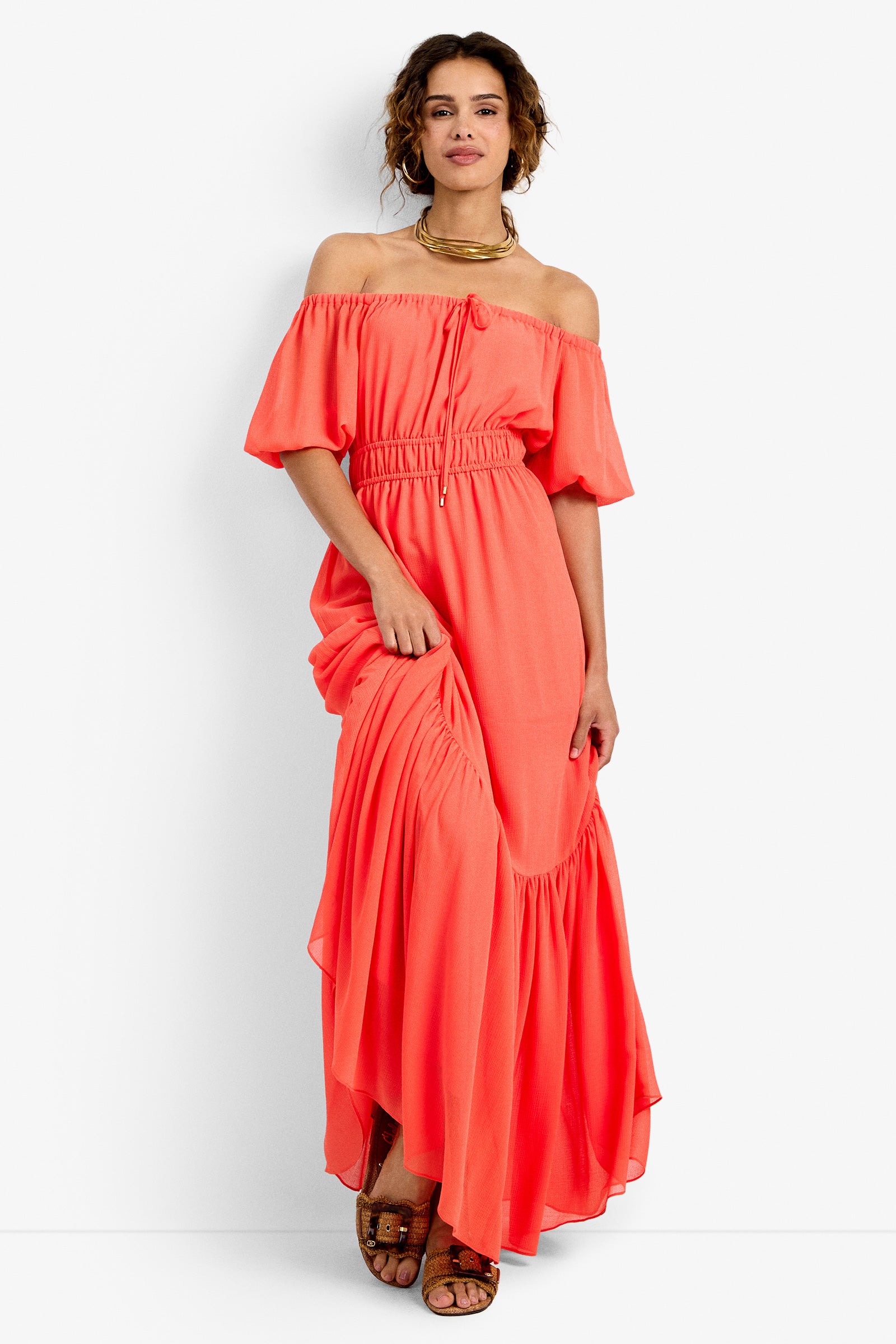 A woman wearing the West Coast Dress in CALYPSO CORAL—an off-the-shoulder maxi with short sleeves and ruffled hem—stands smiling, holding the skirt. She pairs it with sandals and a gold necklace.