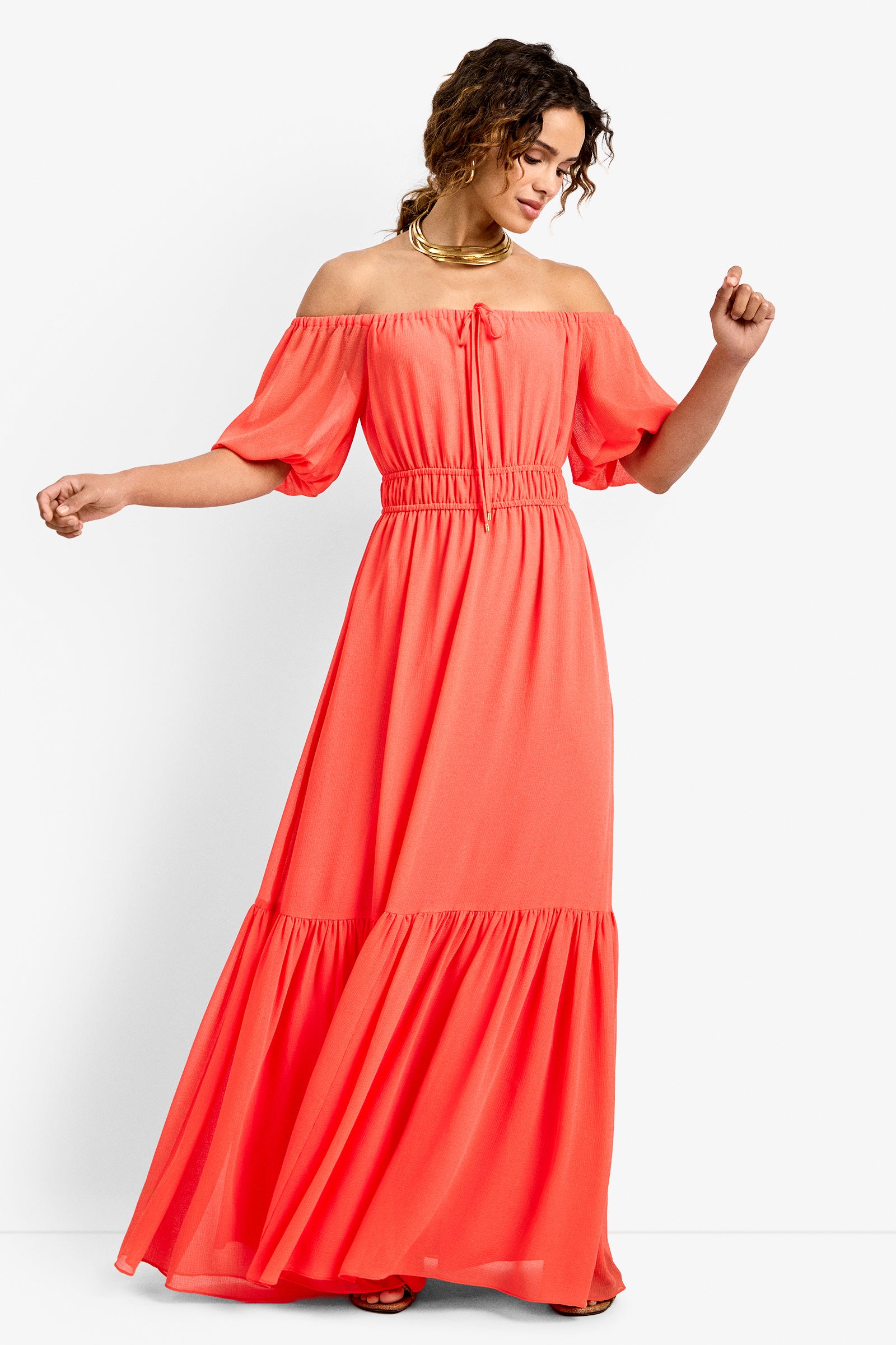 Woman in the West Coast Dress—a coral off-the-shoulder, short-sleeve maxi with a ruffled hem and cinched waist—smiling against a white background with her arms slightly raised.