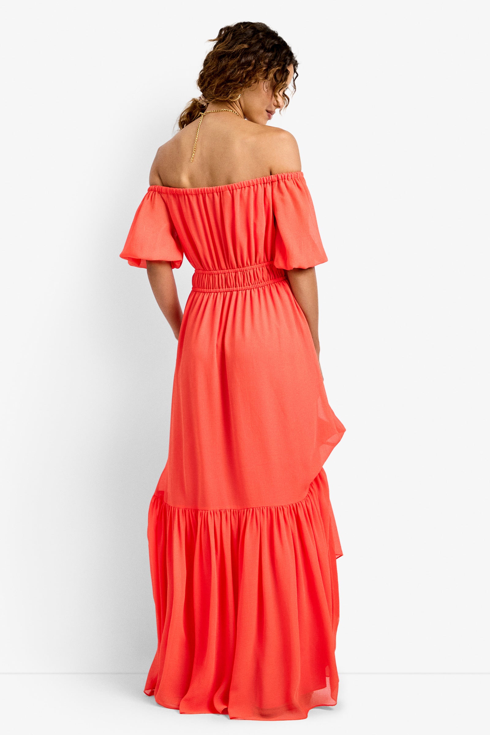 A woman with curly hair wears the West Coast Dress—an off-the-shoulder, coral maxi dress with short sleeves and a ruffled hem—standing and facing away from the camera against a plain white background.