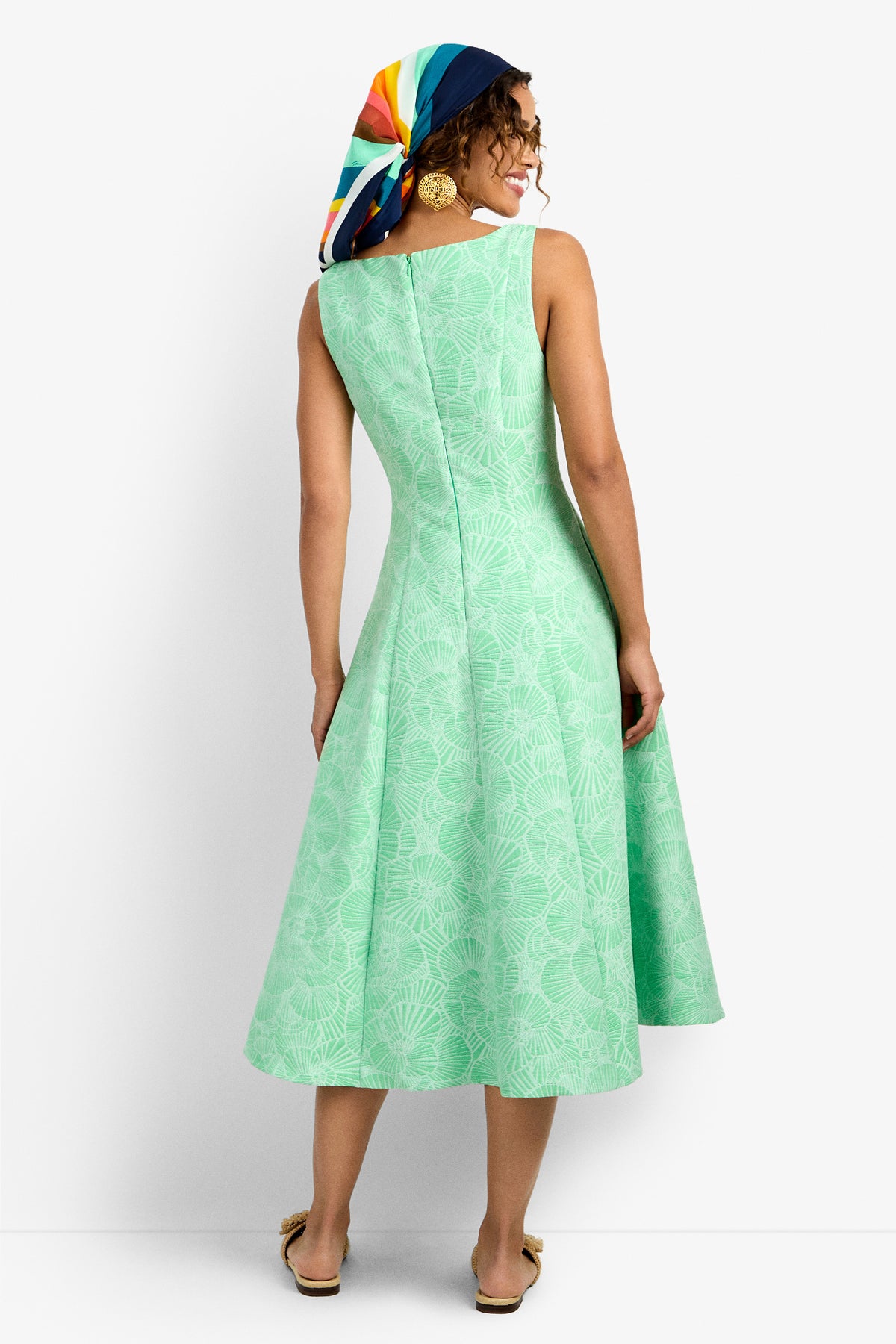 A woman faces away, wearing the Valor 2 Dress—a sleeveless, light green, knee-length dress with a subtle floral pattern. She pairs it with a colorful headscarf, gold earrings, and beige flats.