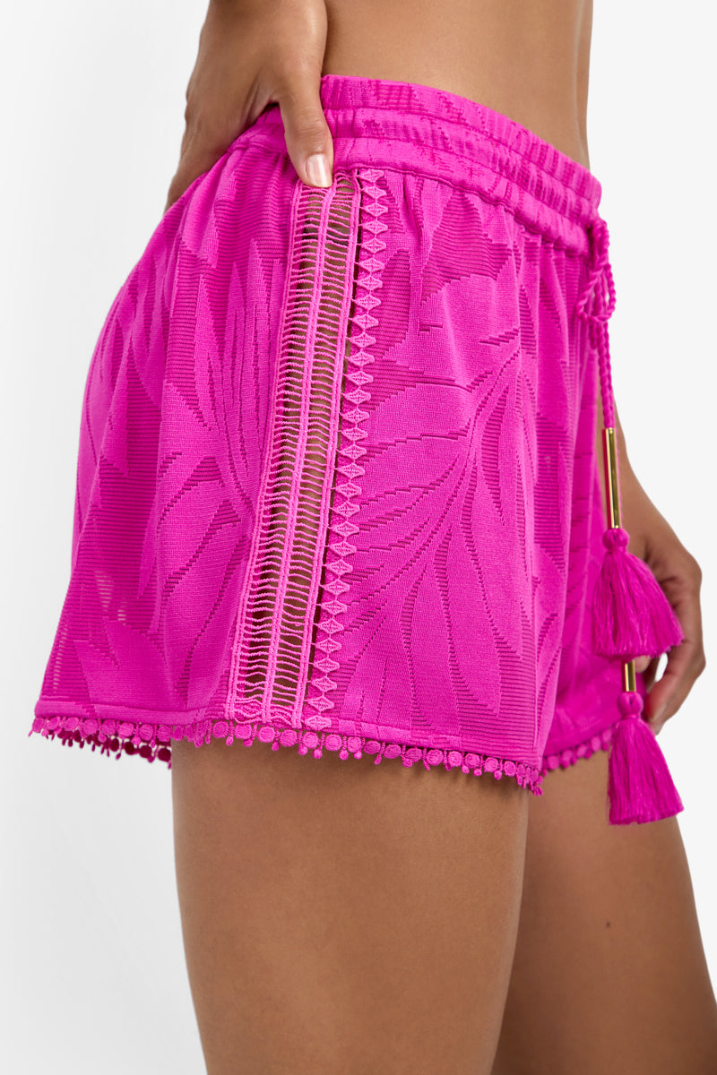 A close-up of a person wearing the Beach Short Cover-Up, featuring bright pink textured fabric with leaf patterns, decorative trim, side lace details, and tassels on the drawstring waistband.