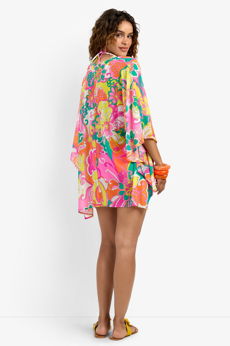 A woman with curly hair stands facing backward, looking over her shoulder and wearing a colorful Kimono Swim Cover-up. She holds orange bangles and completes the look with yellow sandals.