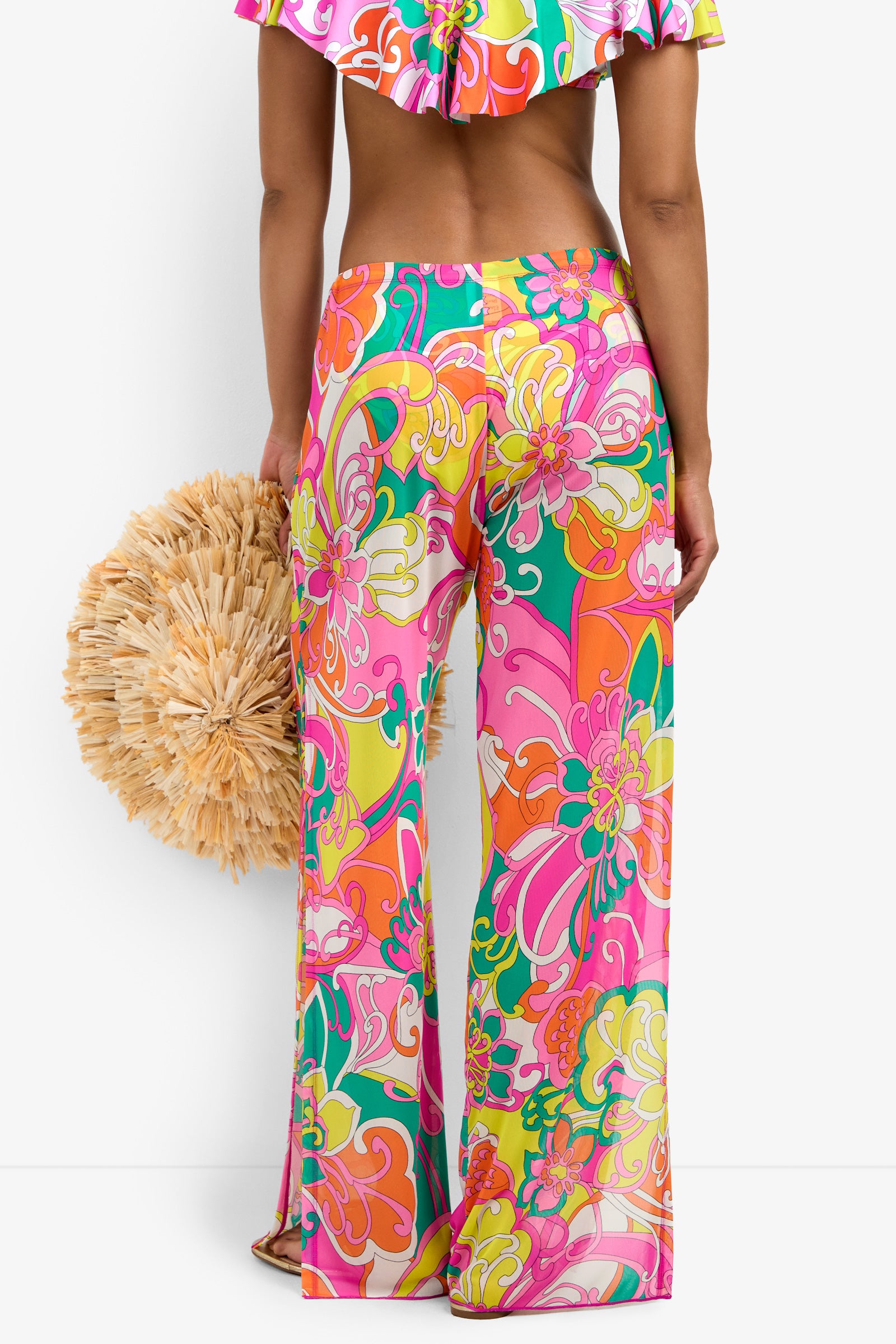 A woman stands facing away, wearing Mesh Swim Cover-up Pants in a vibrant, multicolored floral pattern, paired with a matching crop top and holding a round straw bag.