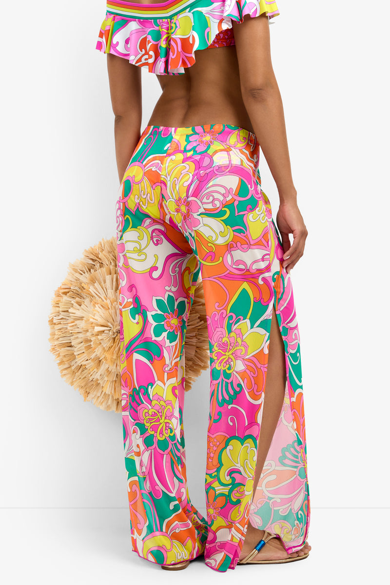 A woman faces away from the camera, wearing Mesh Swim Cover-up Pants with a colorful floral print and high side slits, complemented by a large beige straw bag. She also shows part of a matching off-shoulder top.