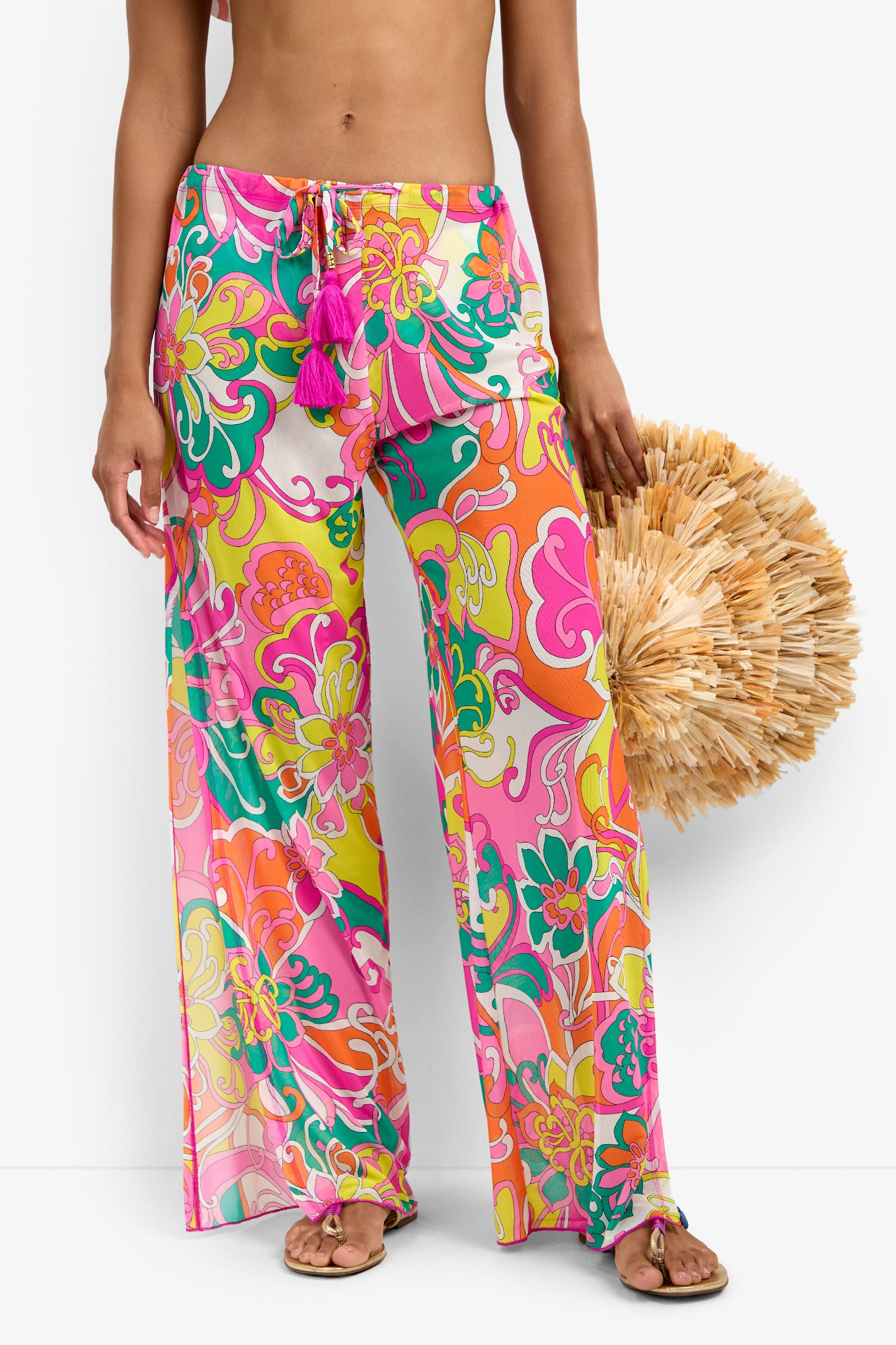 A person wears the Mesh Swim Cover-up Pant—colorful, wide-leg, floral with a pink drawstring and tassels—barefoot, holding a large round straw bag. The image is cropped at the shoulders.