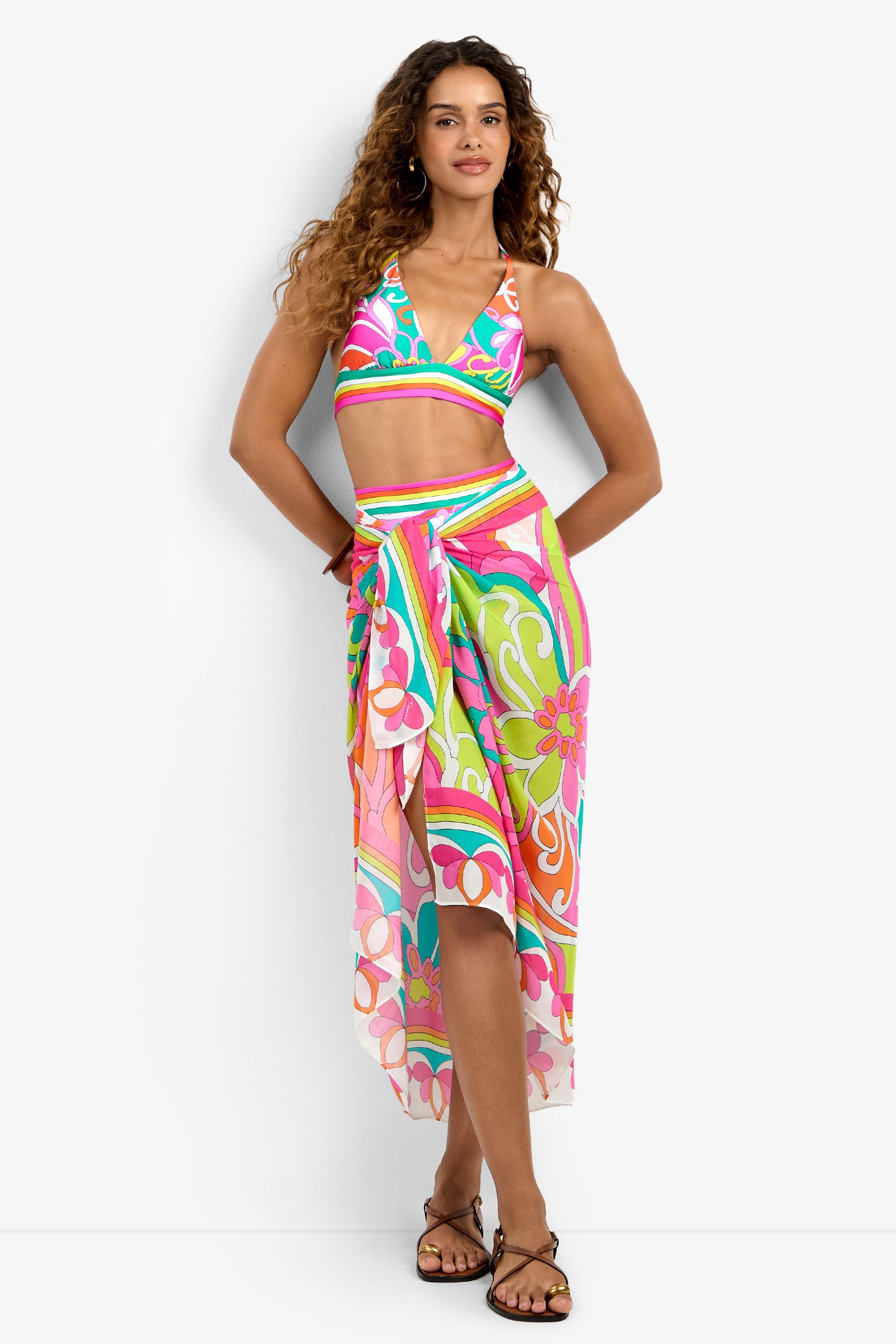 A woman with long curly hair is barefoot on a white background, wearing the Pareo Swim Cover-Up—a colorful tropical-patterned halter crop top and matching wrap skirt featuring pink, green, orange, and white floral designs.
