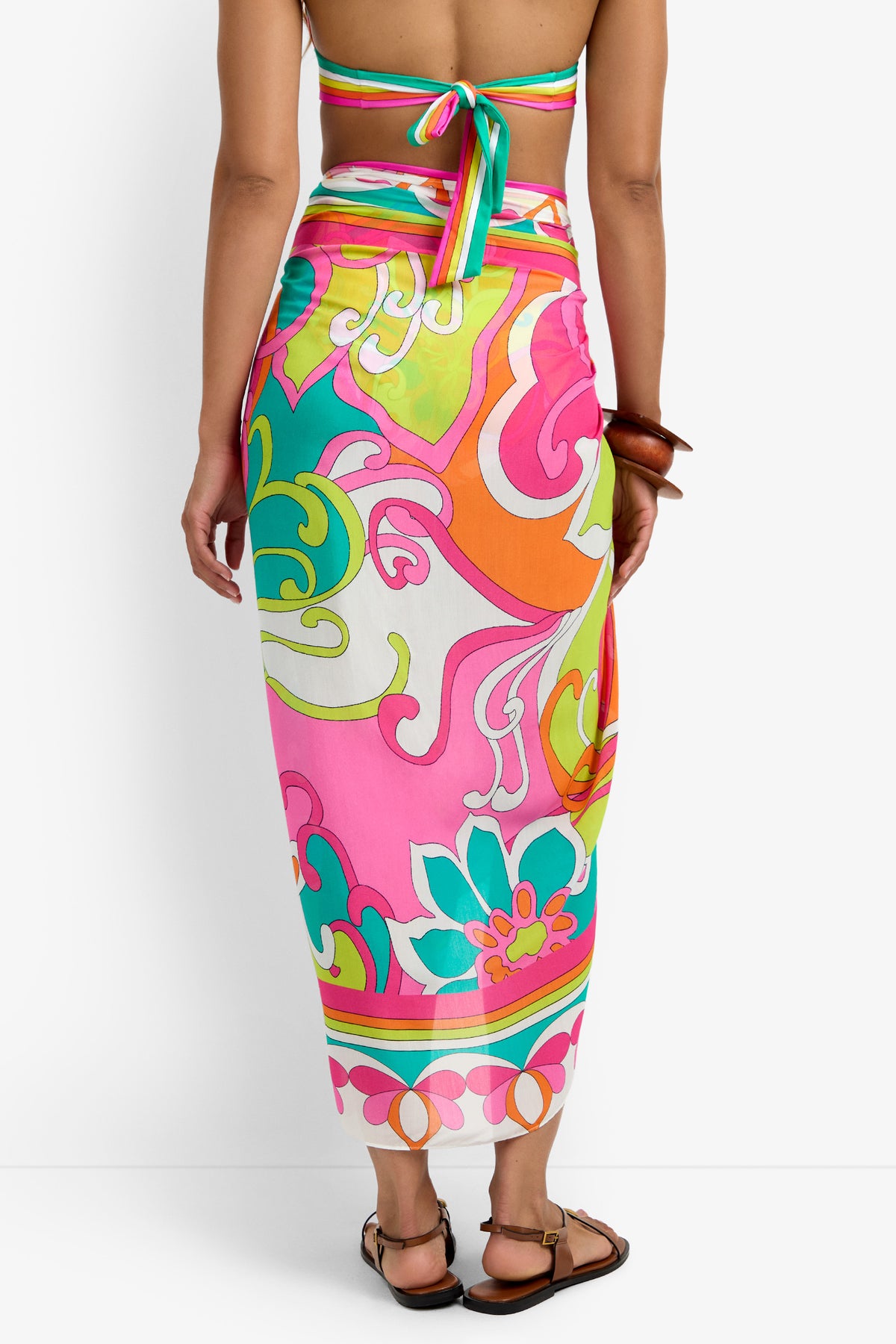 A woman shown from the back wears a brightly colored Pareo Swim Cover-Up with pink, green, orange, and yellow swirls and florals, paired with flat sandals and a halter bikini top.
