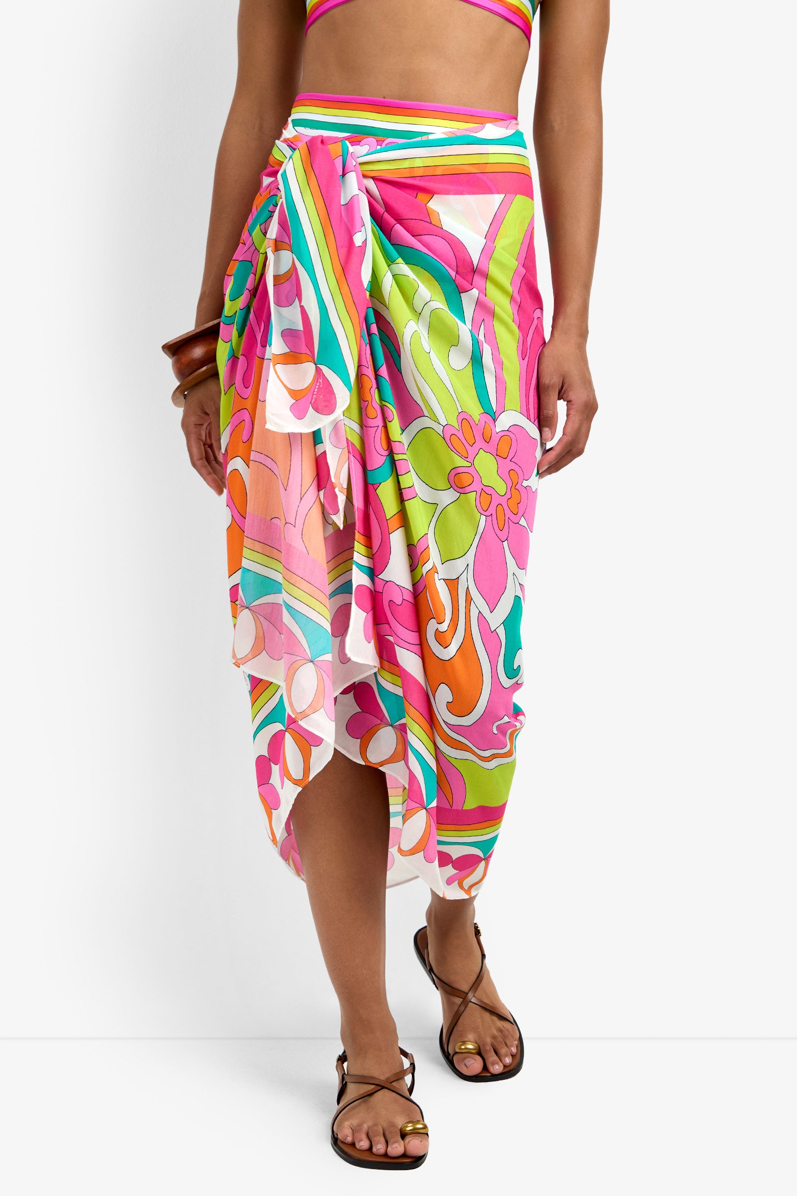 A person wearing the Pareo Swim Cover-Up with a colorful floral pattern in pink, orange, green, and white. The sarong is tied at the side for an asymmetrical hem and paired with a matching top and brown sandals.