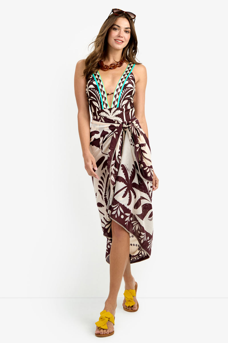A woman stands and smiles against a white background, wearing the Pareo Swim Cover-Up—a sleeveless, deep V-neck dress with a bold brown and cream tropical print—paired with yellow sandals, a chunky necklace, and sunglasses on her head.