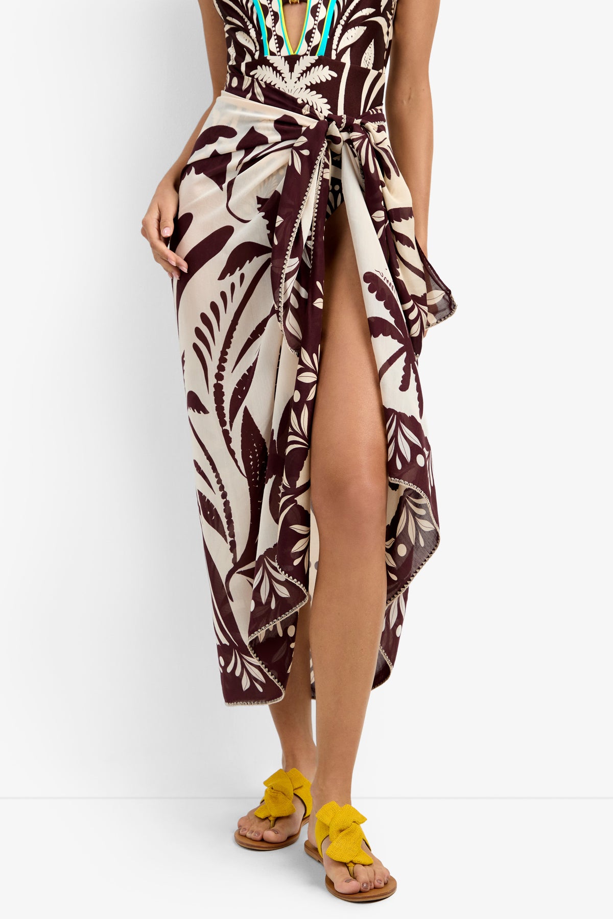 A person wears a Pareo Swim Cover-Up in maroon and beige leaf print, tied at the waist to reveal one leg. They pair it with yellow bow sandals and a colorfully embroidered swimsuit.