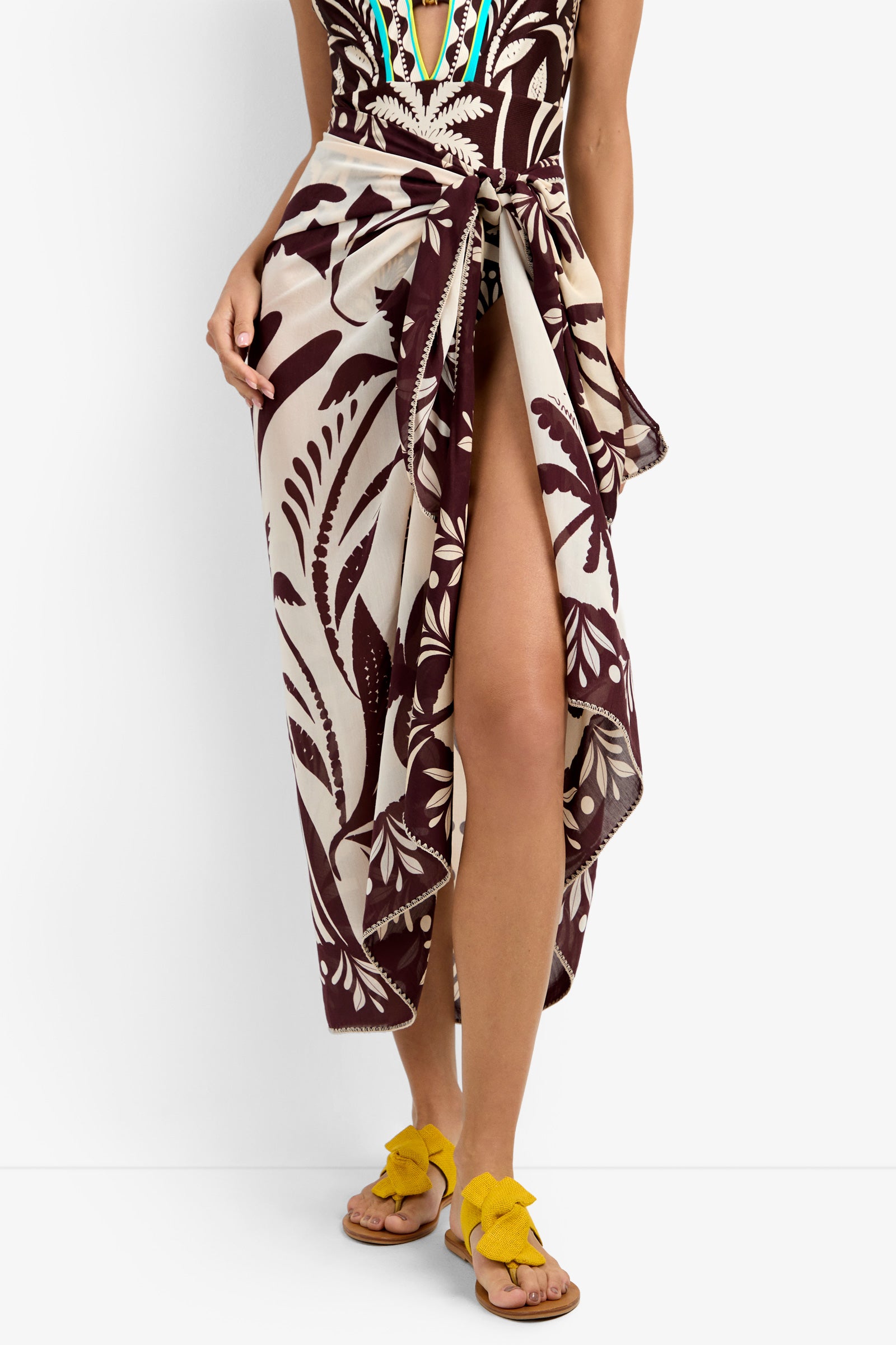 A person wears a Pareo Swim Cover-Up in maroon and beige leaf print, tied at the waist to reveal one leg. They pair it with yellow bow sandals and a colorfully embroidered swimsuit.