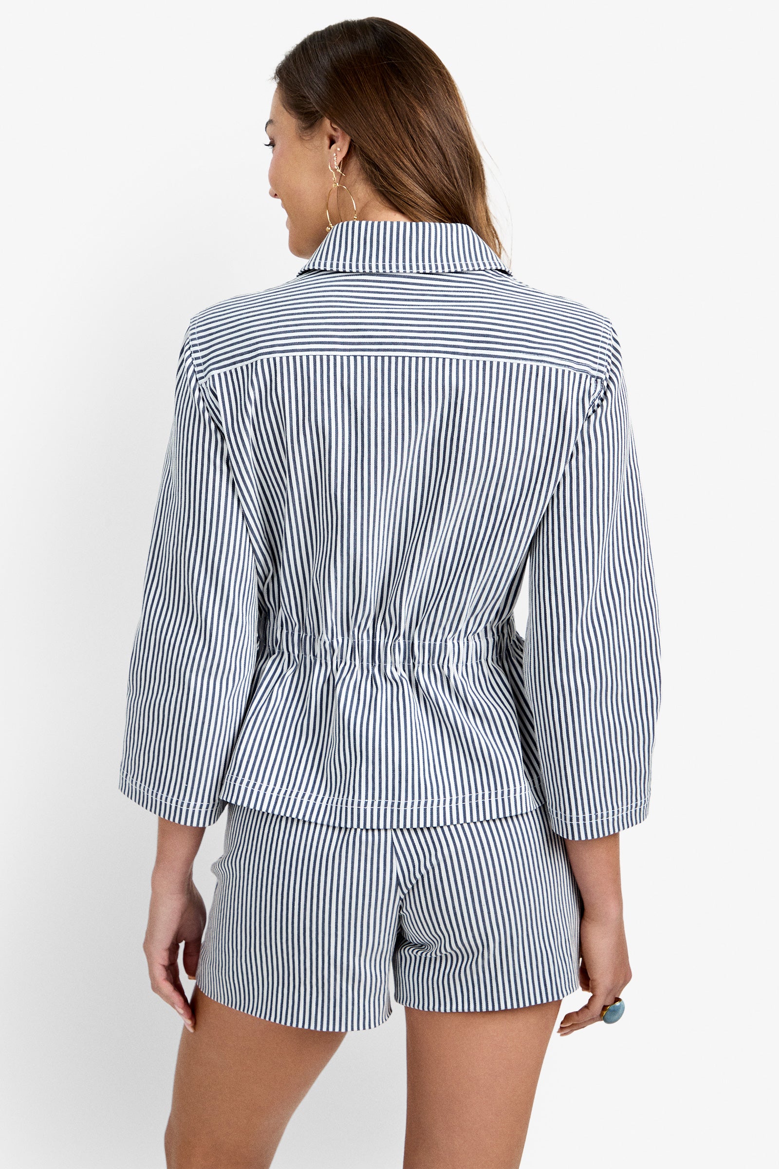 A woman with long brown hair and hoop earrings is seen from the back wearing the Rafferty Jacket, a blue and white vertical striped piece with a cinched waist, paired with matching striped shorts.