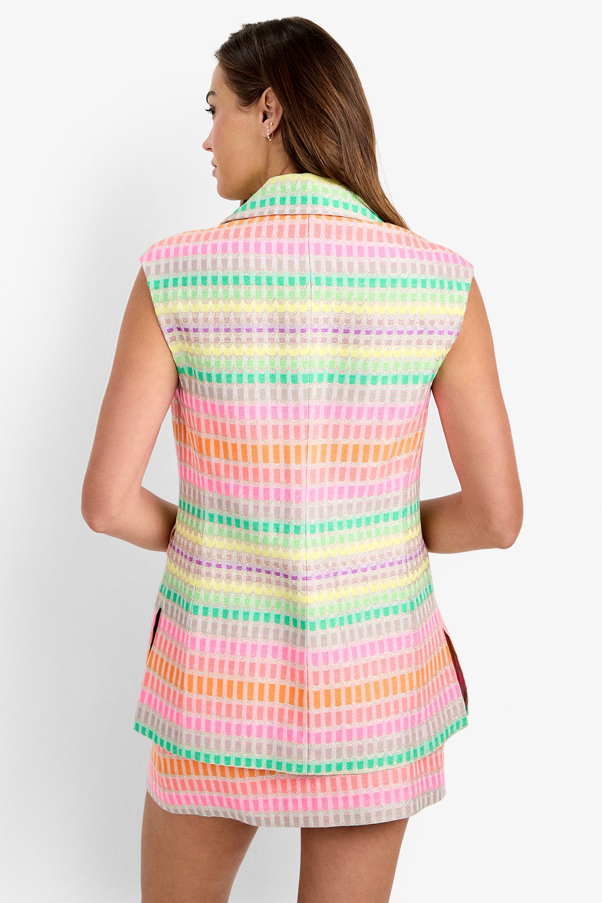 A woman is seen from the back wearing the Boabab Jacket, a sleeveless multicolored vest with a grid pattern in pastel pink, orange, green, and yellow, paired with a matching skirt.