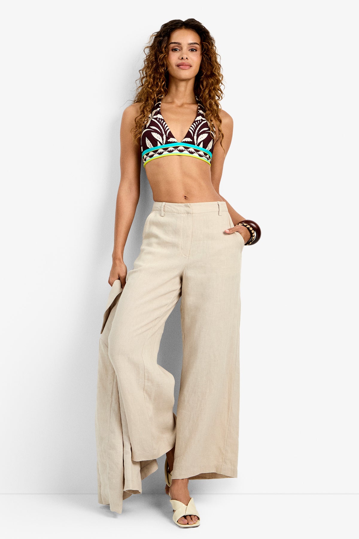 Woman with curly hair stands confidently against a plain light background, wearing the Tonga Pant in Beachy Beige, paired with a patterned halter crop top, cream sandals, and bracelets.