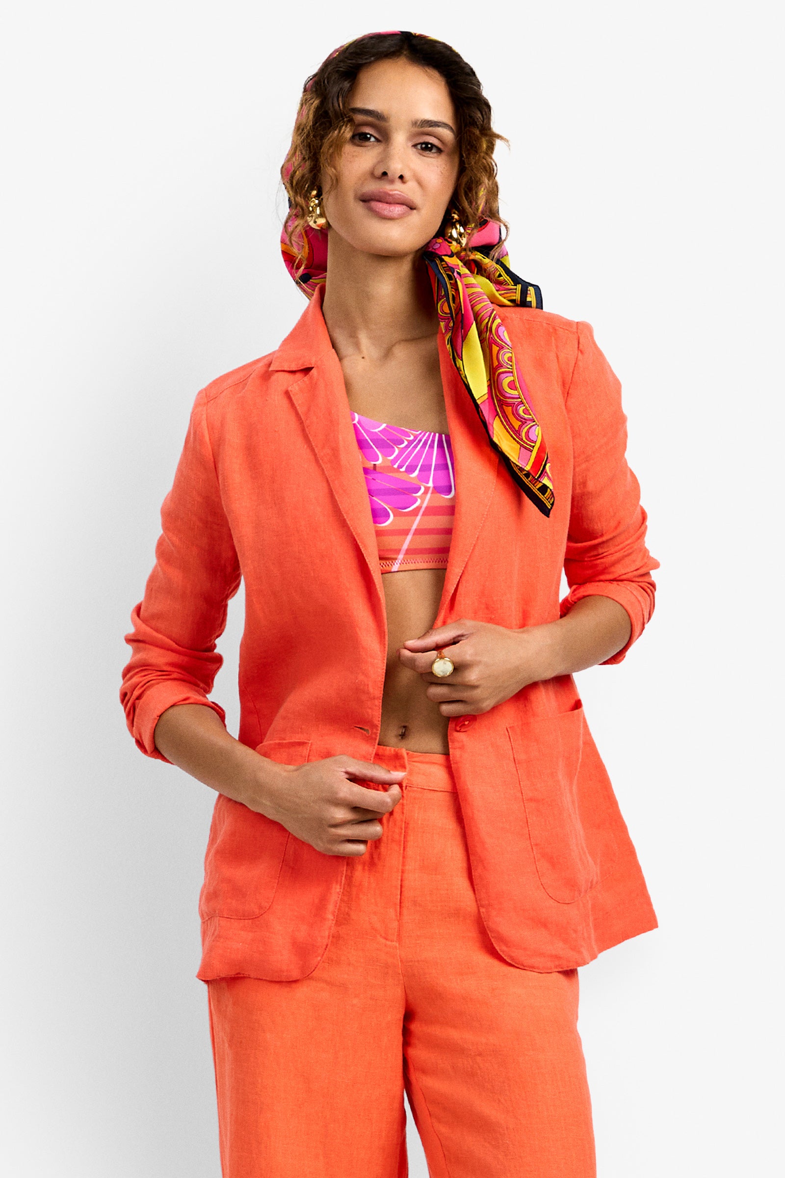 A woman stands confidently against a plain white background, wearing the Azurine Blazer in orange with matching pants, a colorful patterned hair scarf, and a pink patterned crop top.