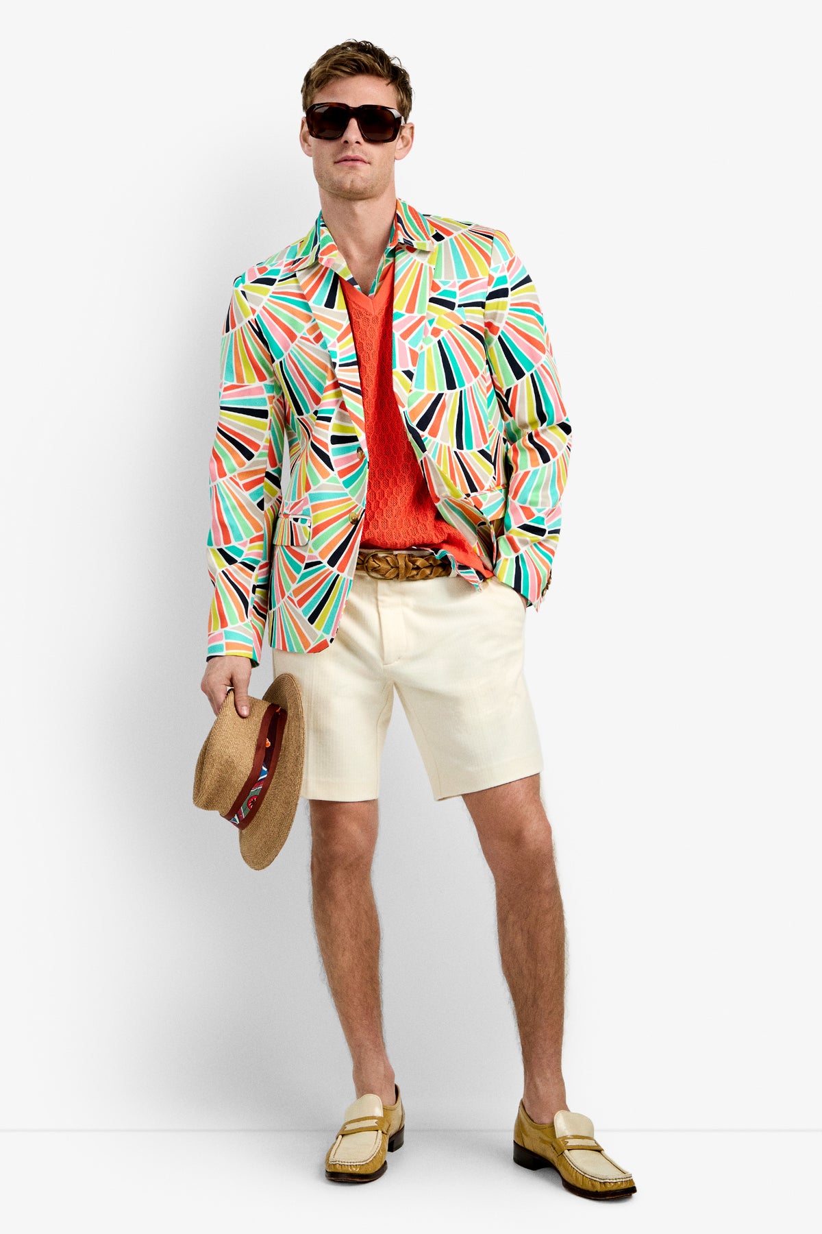 A man in the THURSTON BLAZER (MULTI) with sunglasses, an orange shirt, white shorts, beige loafers, and holding a straw hat stands against a plain white background.