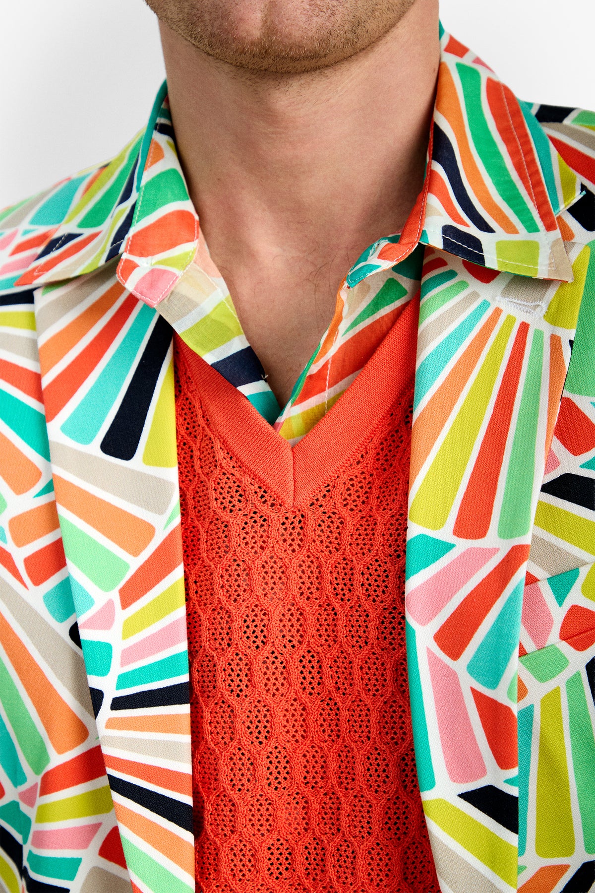 Wearing the THURSTON BLAZER, a colorful geometric-patterned jacket, layered over an orange textured V-neck sweater and shirt. Only the lower face and neck are shown.