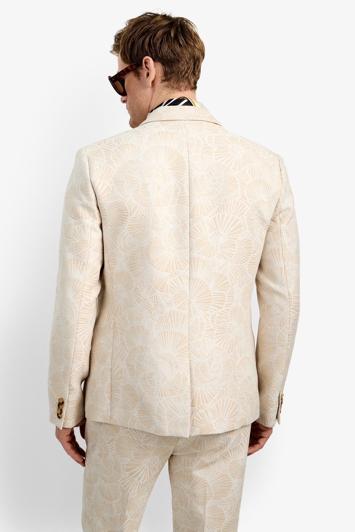 A person with light brown hair and sunglasses is seen from behind wearing the cream-colored Thurtson Blazer, which features a subtle shell-like pattern and gold buttons on the sleeves.