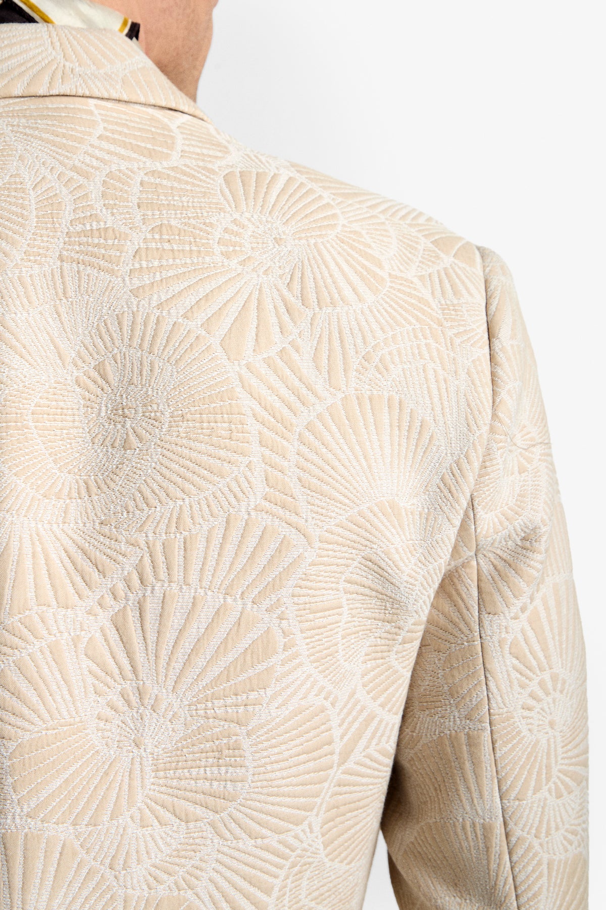 Close-up of the Thurtson Blazer’s back in beige, featuring an intricate, textured pattern like overlapping seashells or fans. A glimpse of the wearer’s neck and shoulder is shown against a plain white background.